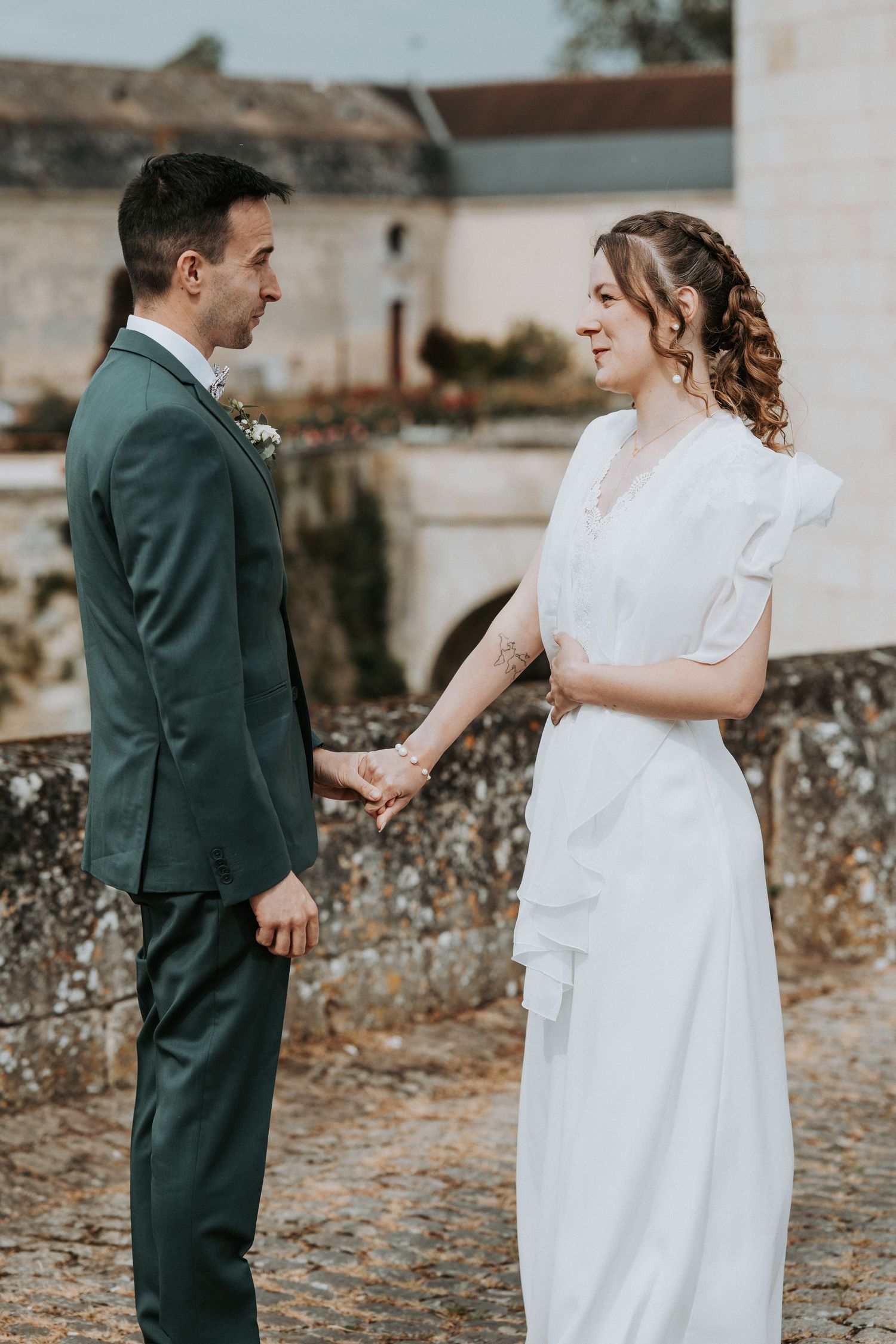 A bride and groom hold hands outdoors, smiling at each other, she in white dress, he in green suit.