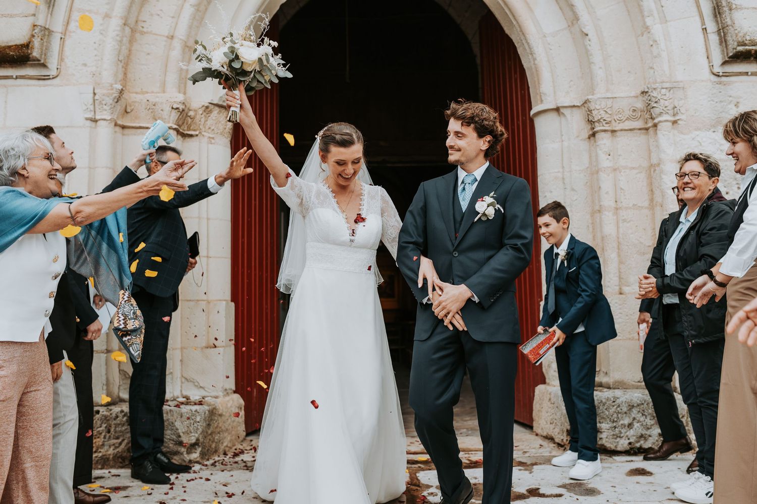 Wedding party celebrates with confetti and laughter outside a historic stone church archway.