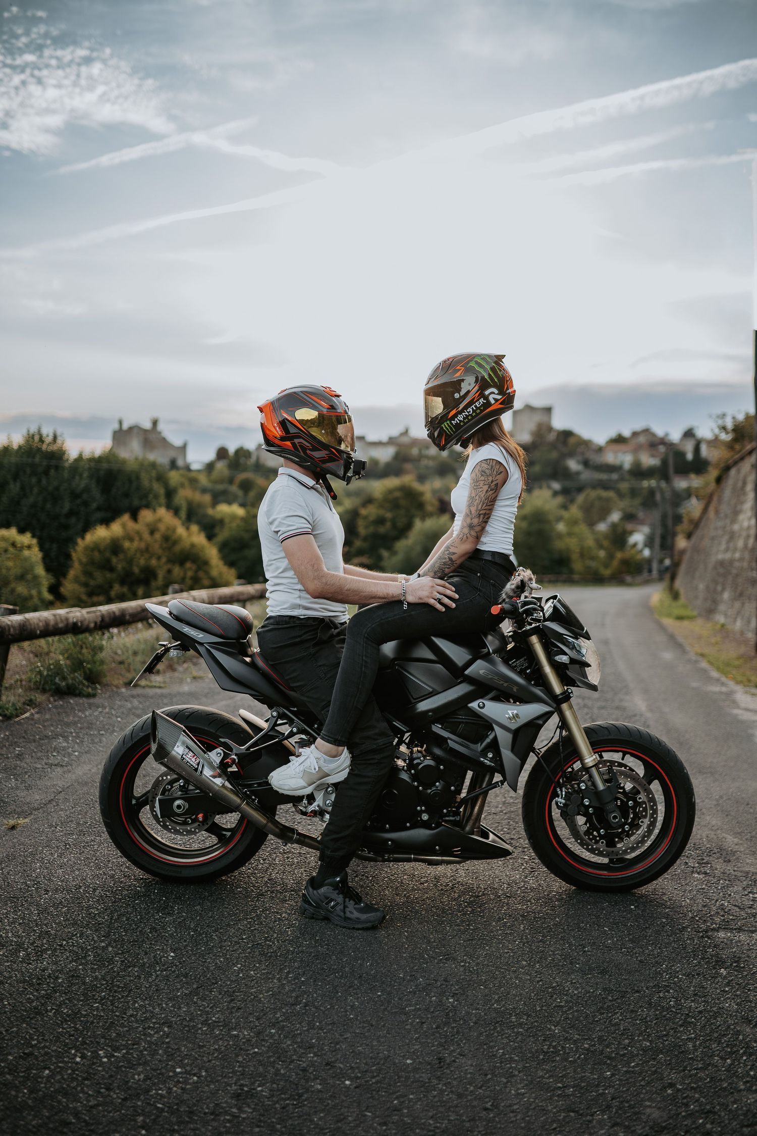 Two motorcycle riders in protective gear sit together on a black sport bike against a scenic backdrop with houses.