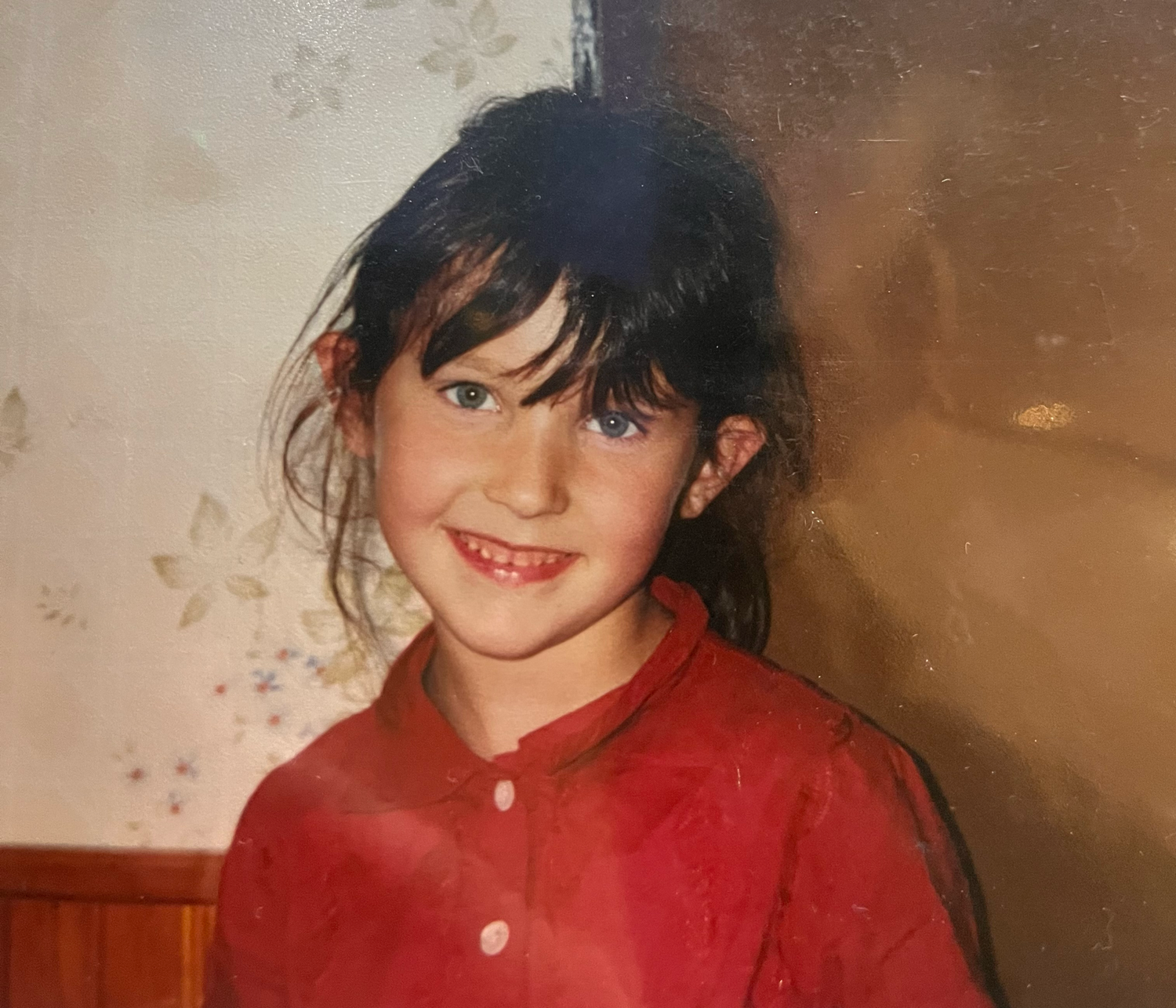 Young girl with dark hair smiling in a red top, vintage childhood photo with floral wallpaper background.