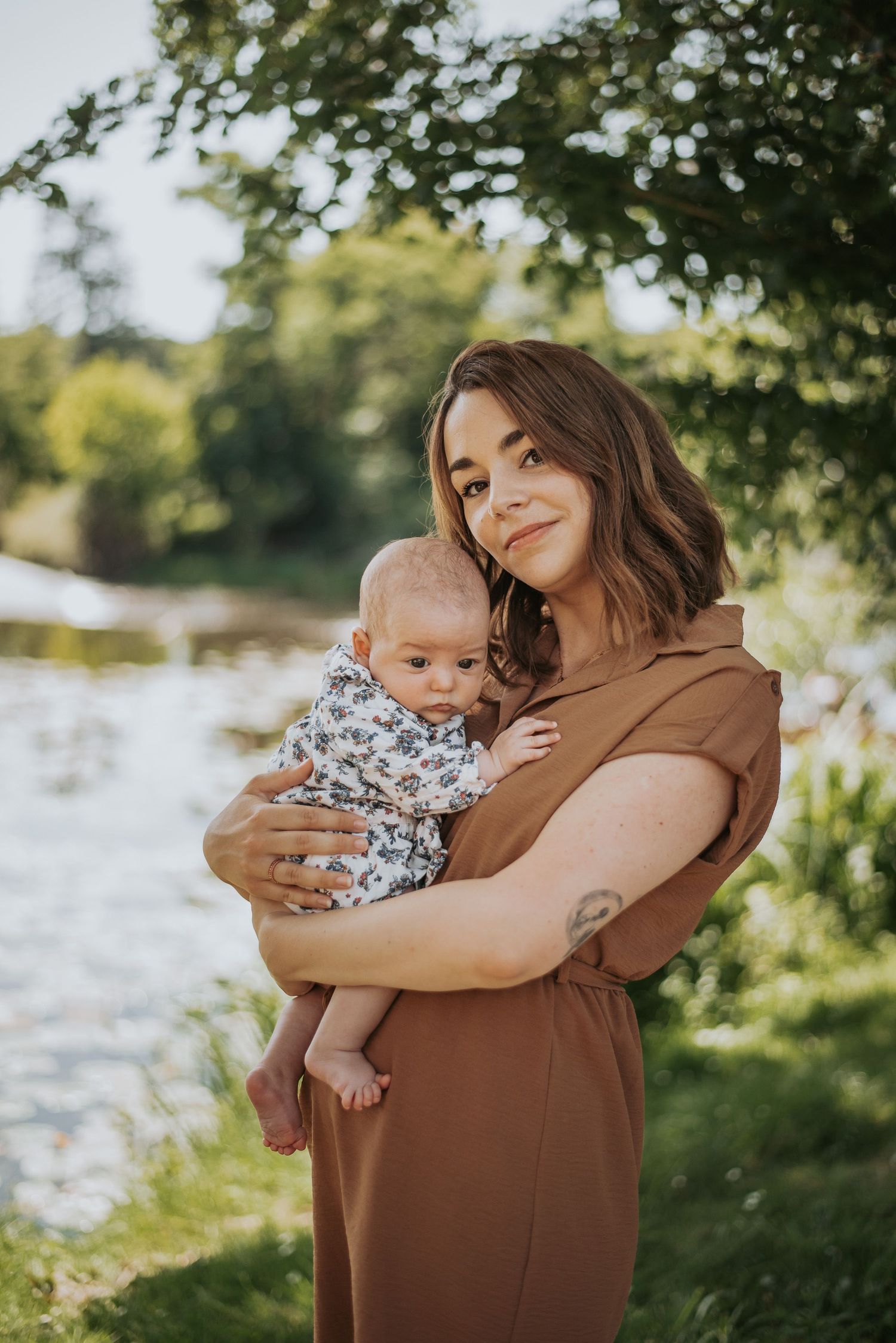 A parent hugs and comforts a smiling baby wearing a floral onesie by a sunlit lake on a summer day.