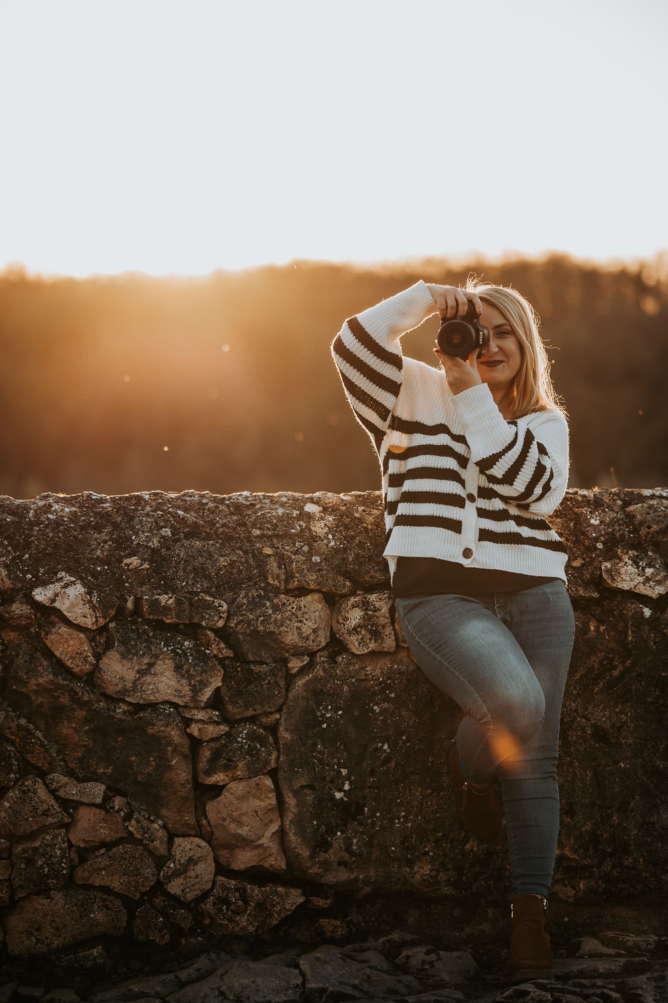 Woman in striped sweater and jeans photographing with camera against stone wall in golden sunset light.