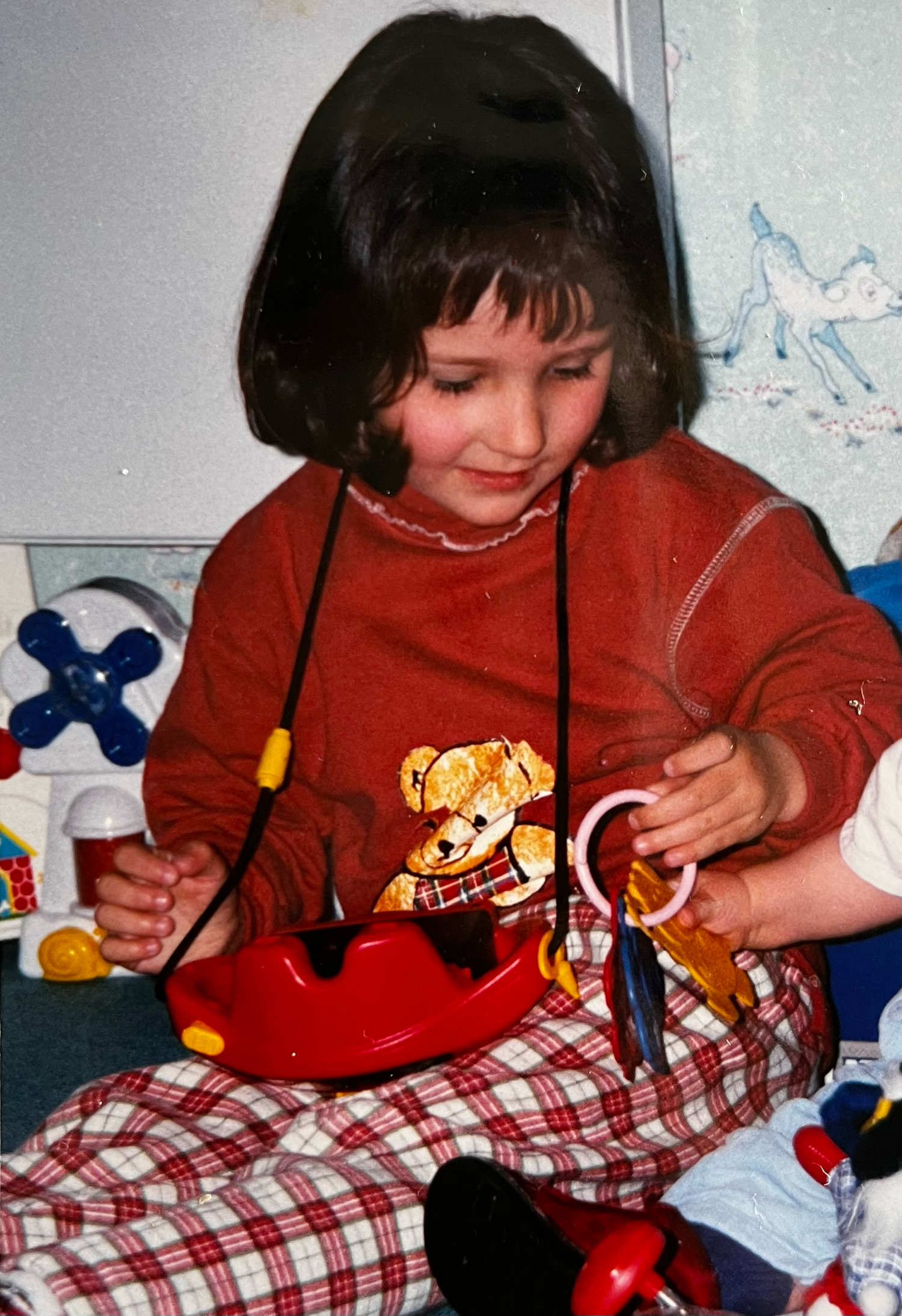 A young child in a red sweater with a puppy design plays with toys while wearing plaid pants.