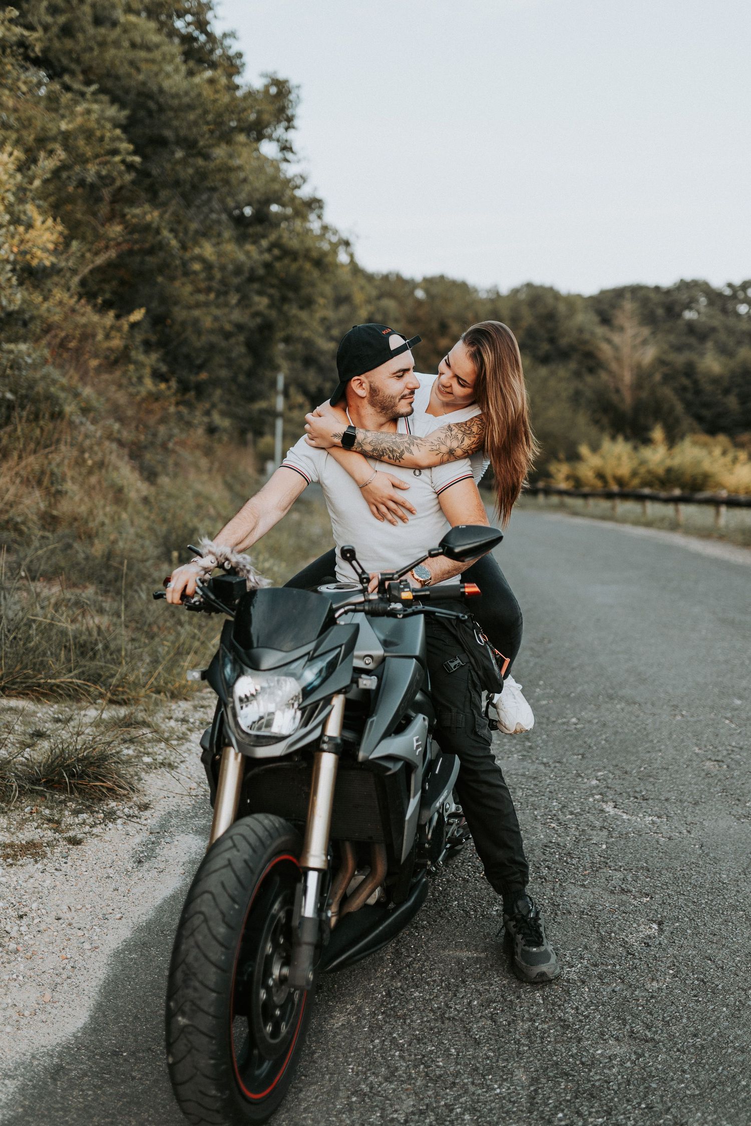 Couple posing with a black sports motorcycle on a gravel road during an outdoor photo session.