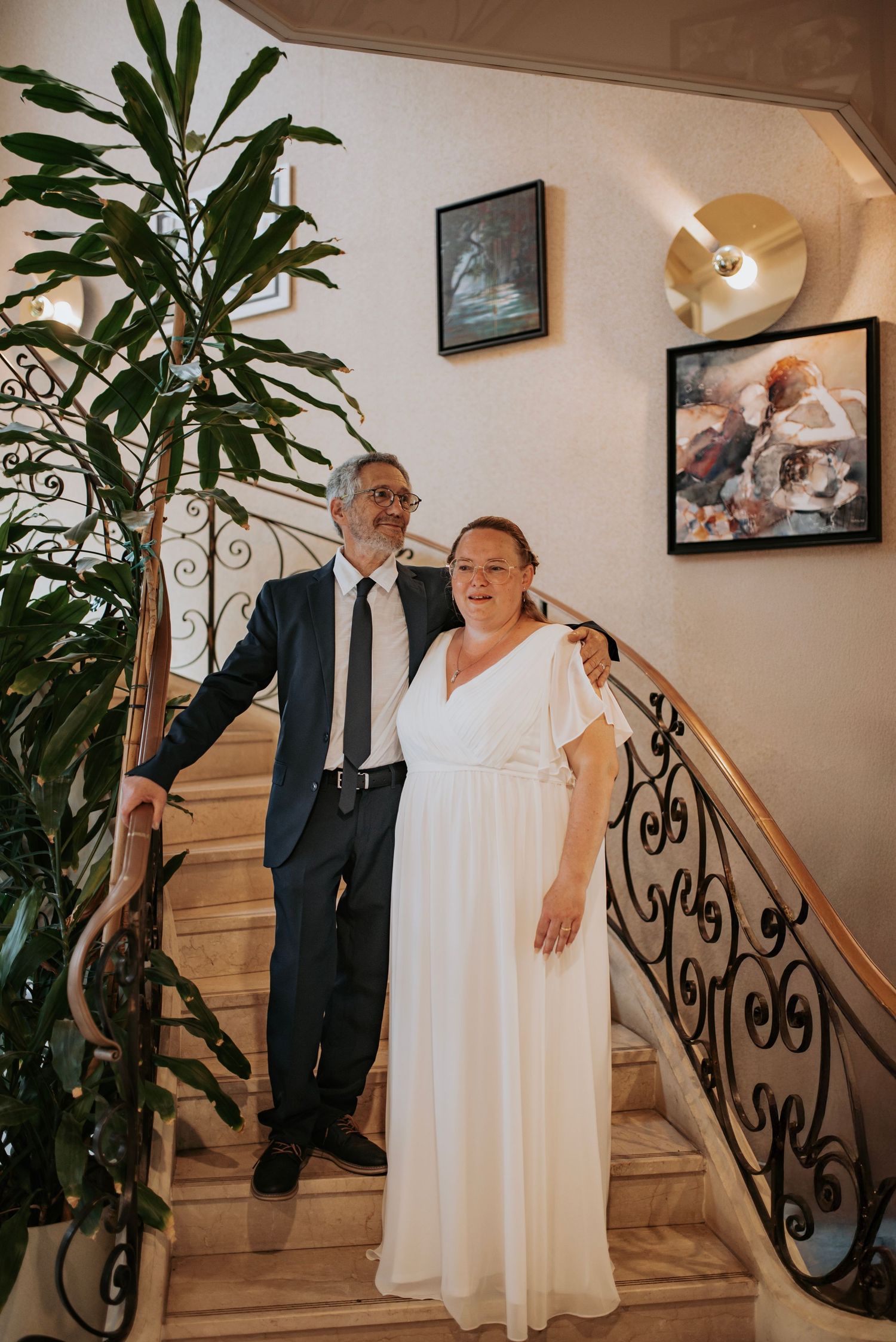 A couple shares joyful moments on an elegant staircase with wrought iron railings and indoor plants during a wedding celebration.