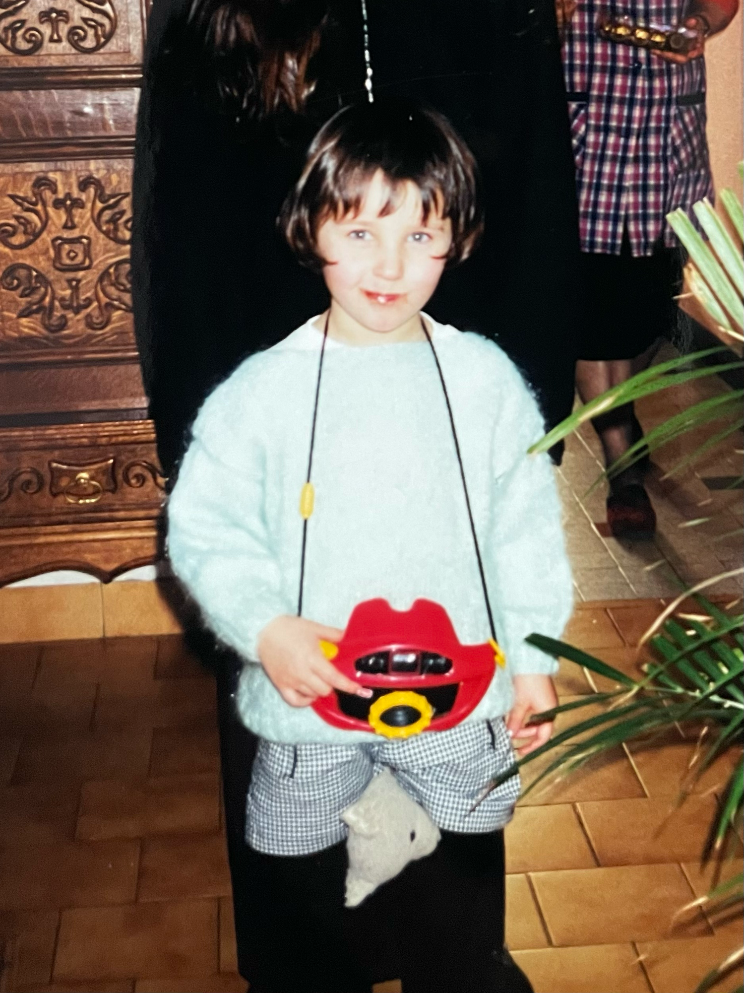 A young child holding a red toy camera with a yellow lens, wearing a light blue sweater and checkered pants indoors.