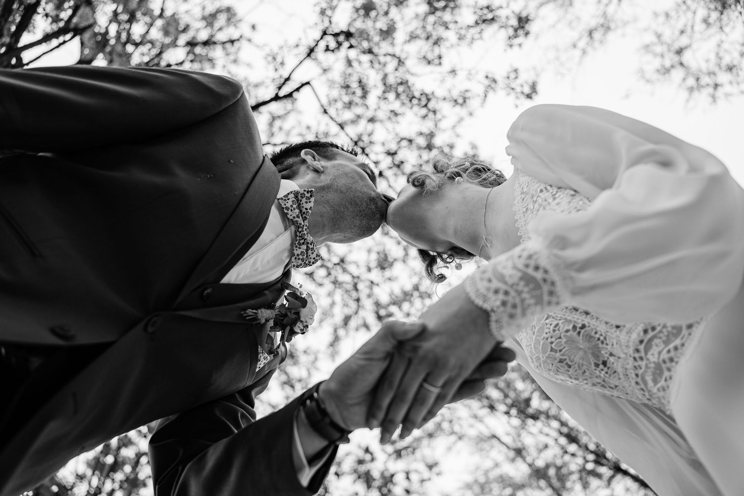 Black and white photo of a bride and groom kissing outdoors, captured from below with trees in the background.
