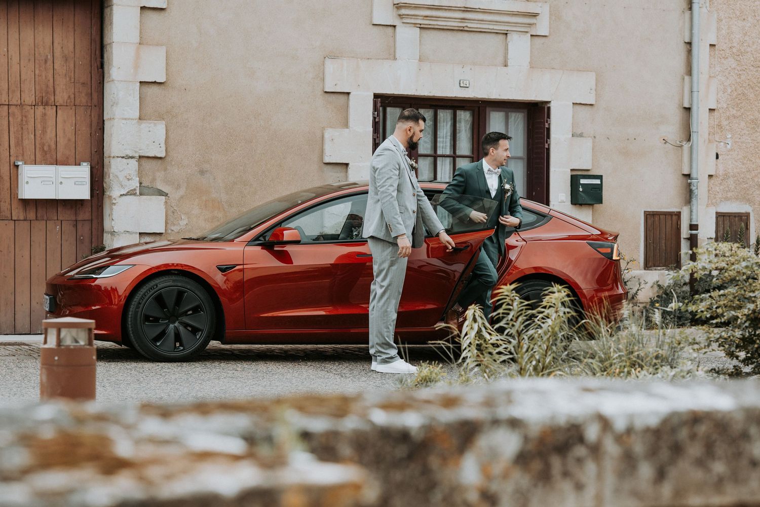 Two men in formal attire stand beside a red Tesla Model 3 outside a stone building.