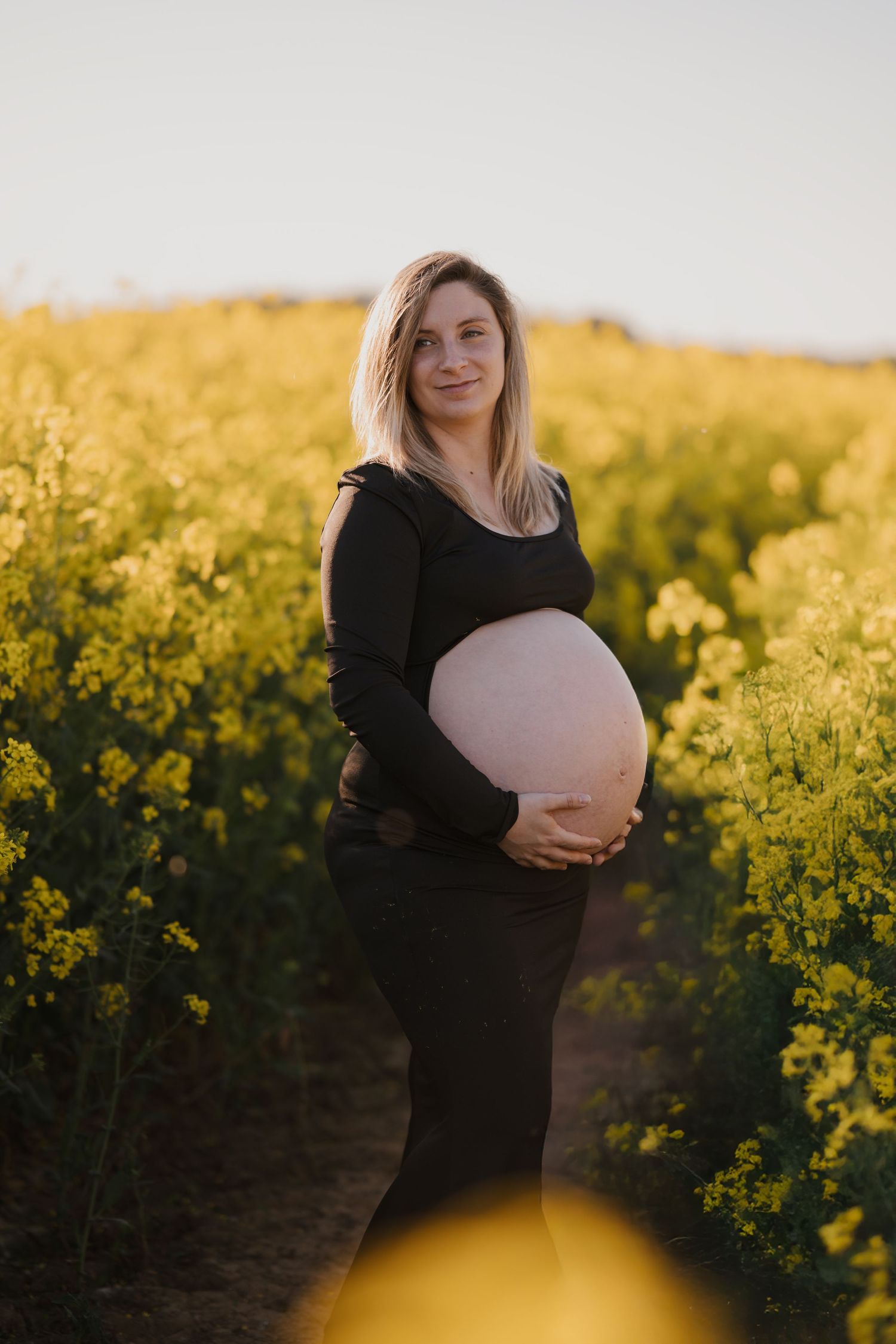 Pregnant woman in black outfit cradling baby bump in golden rapeseed field at sunset.