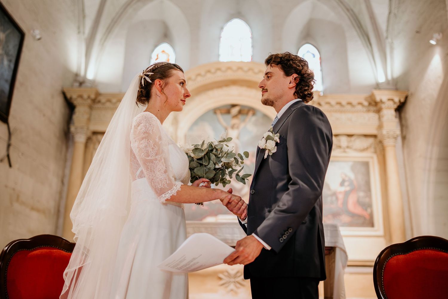 Bride and groom exchange vows holding hands at a church altar, bride in white lace gown with veil, groom in gray suit.