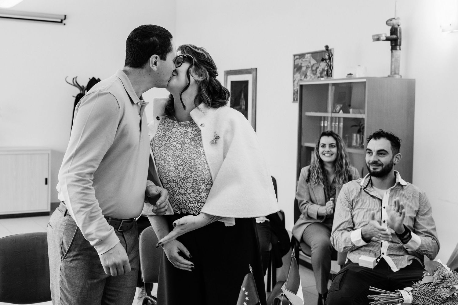 Couple sharing a kiss at an indoor celebration while guests smile and watch in black and white photo.
