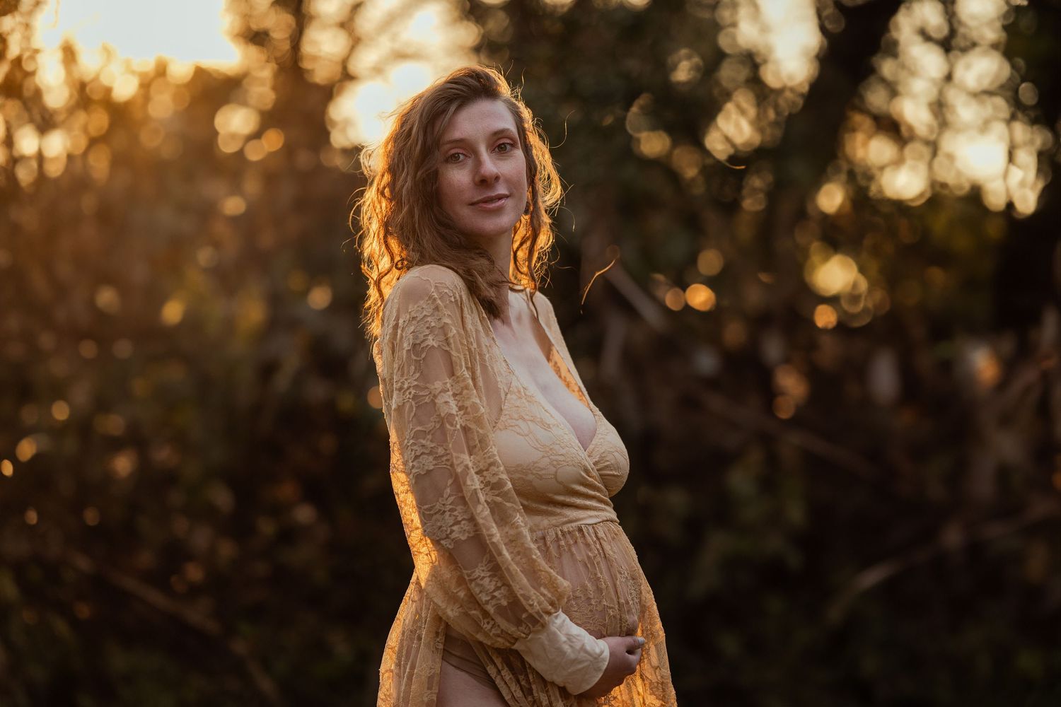 Pregnant woman in golden lace dress glowing in sunset light among trees.