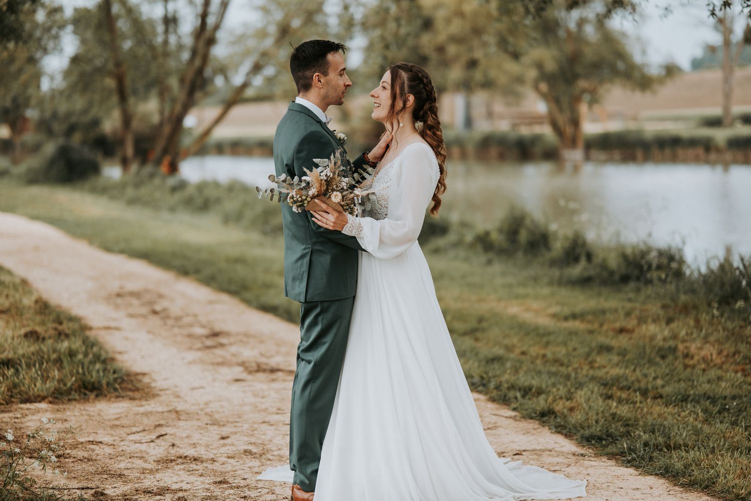 A bride and groom share a romantic moment by a lakeside, she in a white gown, he in a teal suit holding a bouquet.