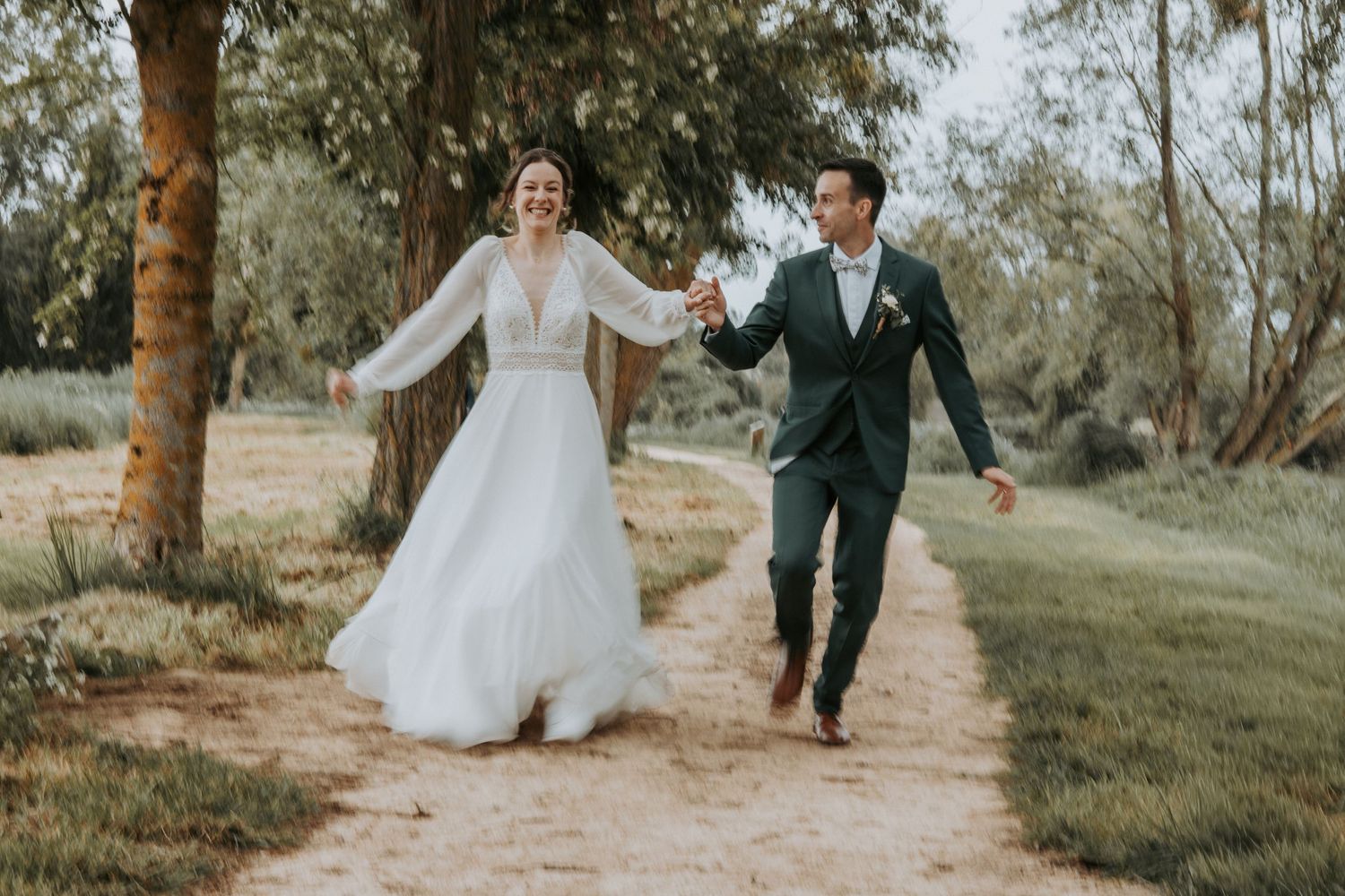 Happy newlyweds holding hands and running on a dirt path surrounded by lush green trees on their wedding day.
