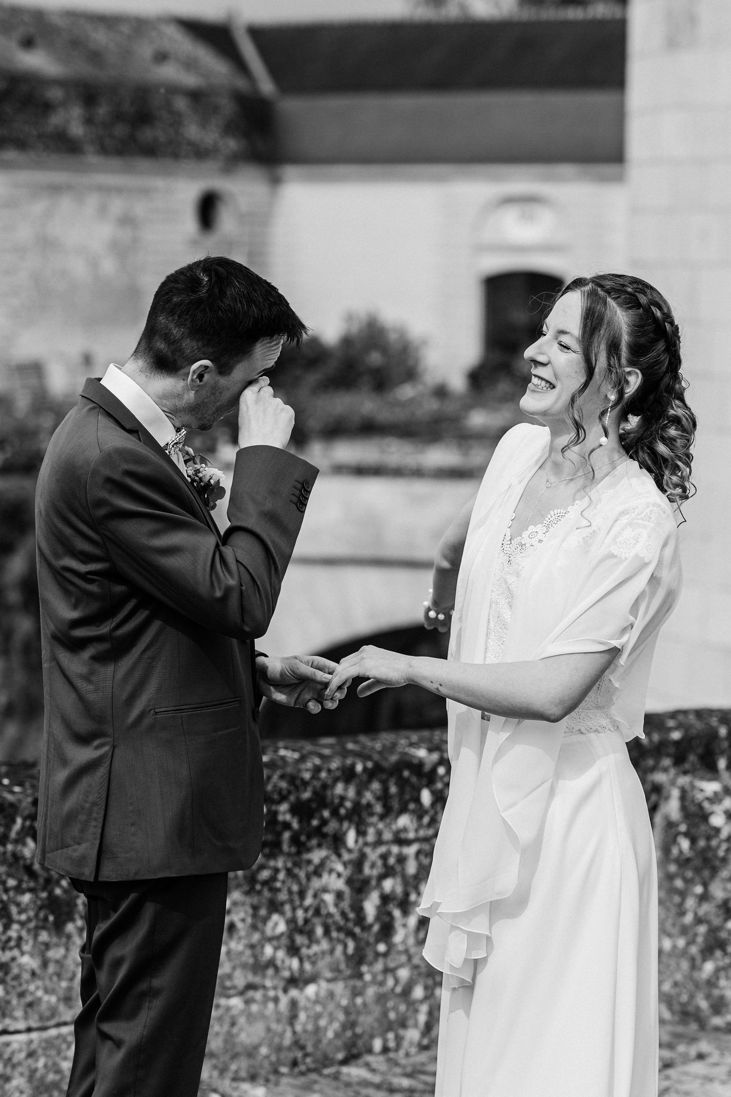 Emotional groom wipes tears while laughing bride holds his hands during outdoor wedding vows in black and white.