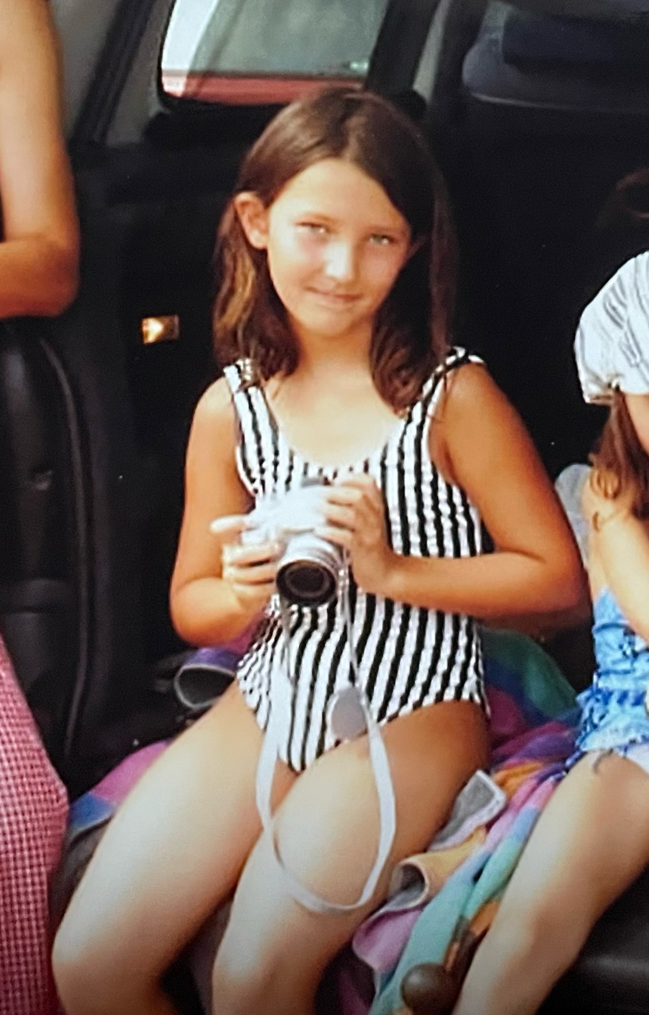 Young girl in black and white striped swimsuit holding a white camera while sitting in a vehicle.