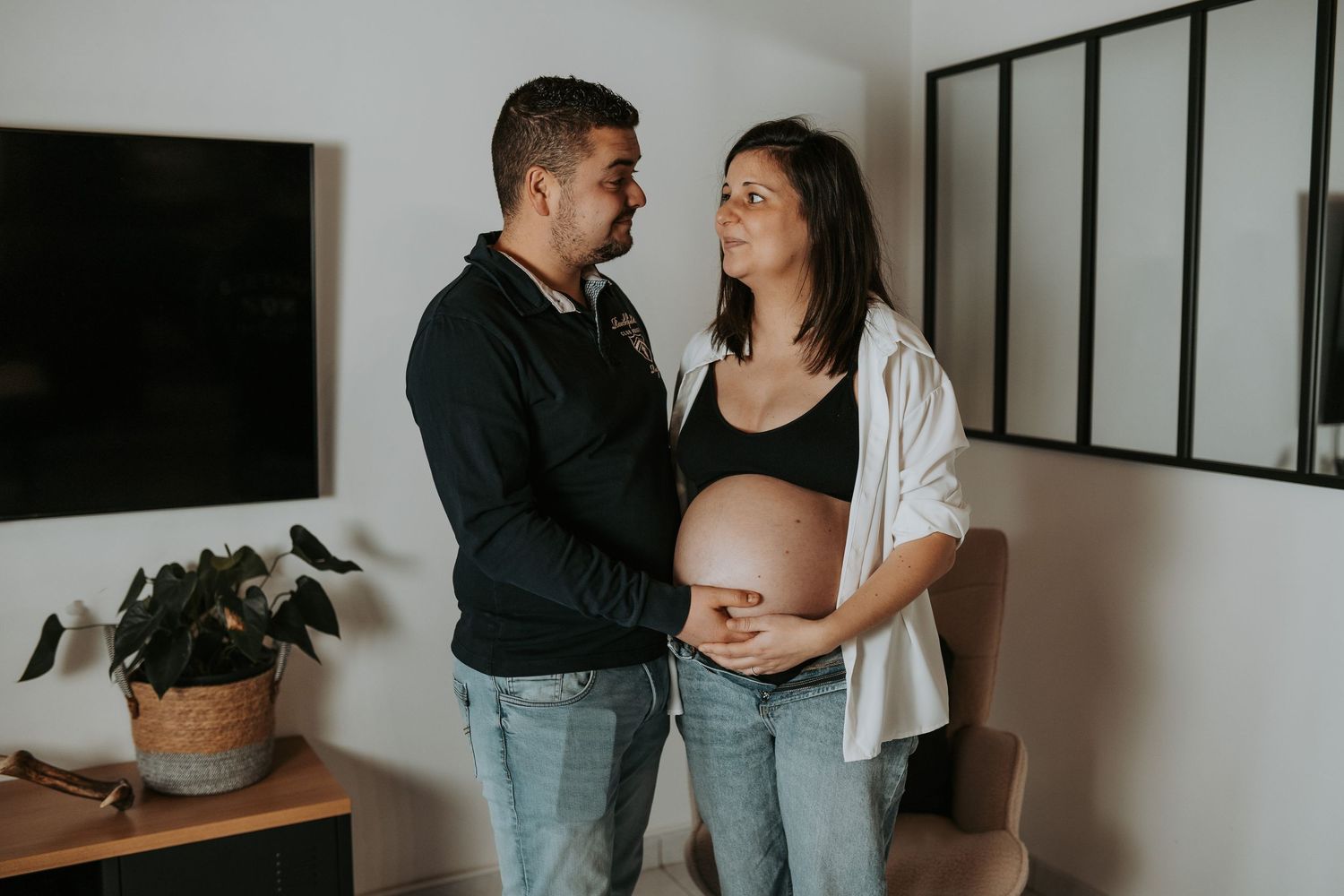 A couple shares an intimate moment at home while expecting a baby, with the pregnant person wearing a white cardigan.