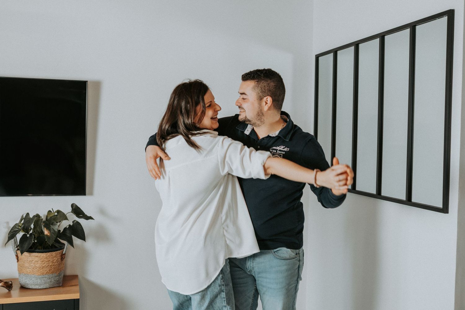 A couple dancing together in a bright modern living room with minimalist decor and a potted plant.