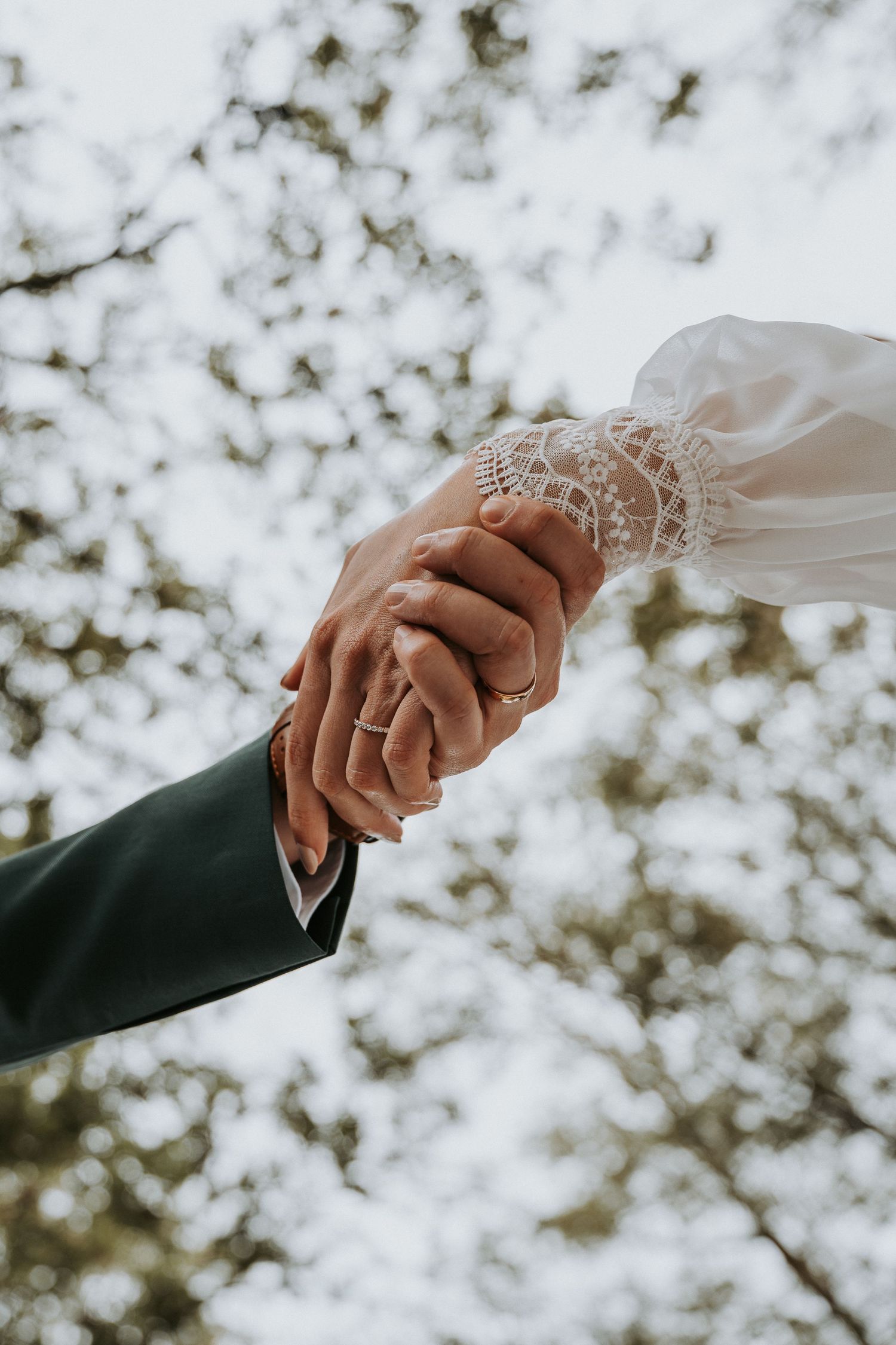 Bride and groom holding hands outdoors, showing wedding rings, lace bridal sleeve and dark suit sleeve against blurred trees.