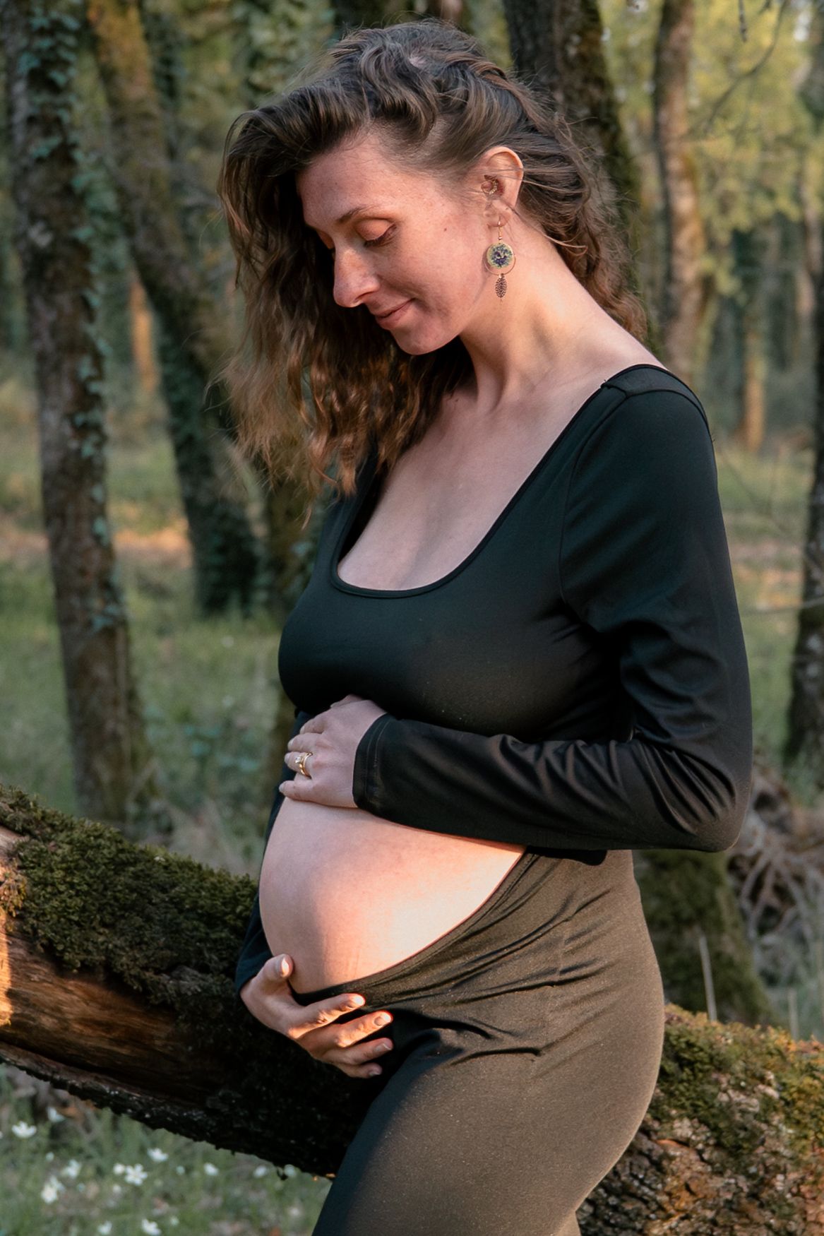 Pregnant woman in black top cradling baby bump outdoors in forest setting during golden hour.