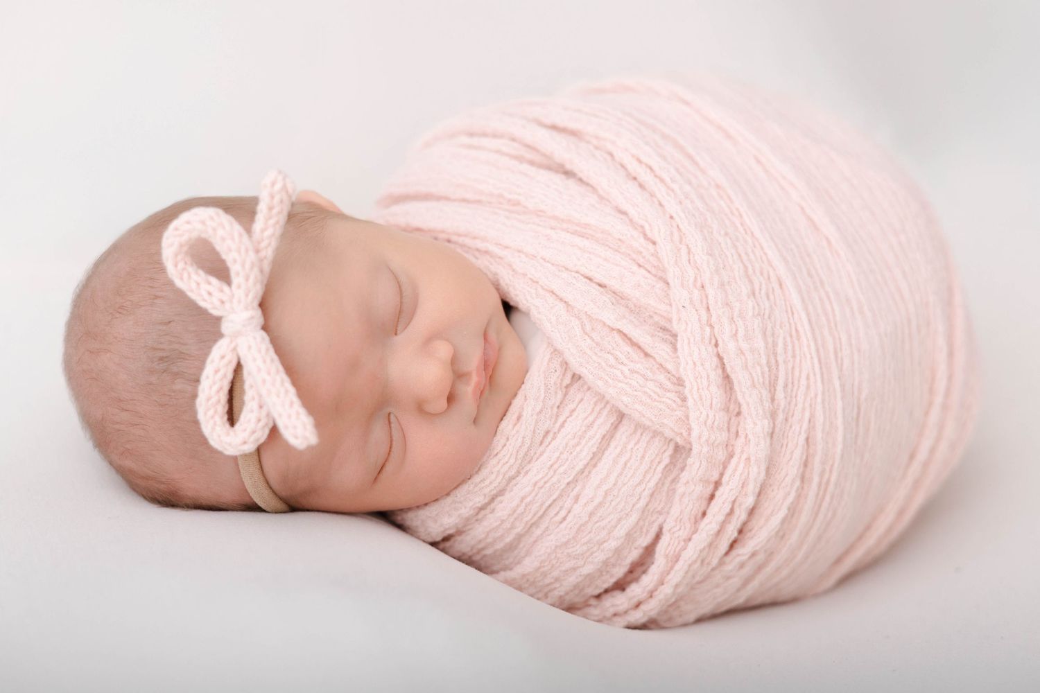 Newborn baby wrapped in delicate pink fabric rests serenely on white backdrop during professional photo session.