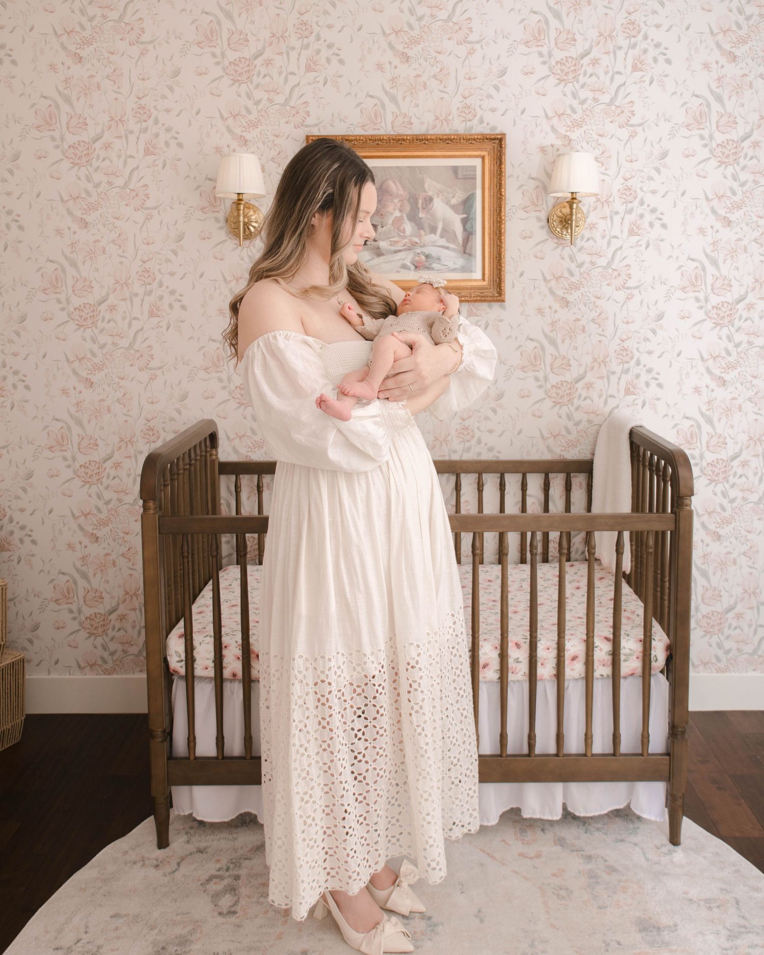 A nursery scene with a woman in white dress holding a baby beside a wooden crib.