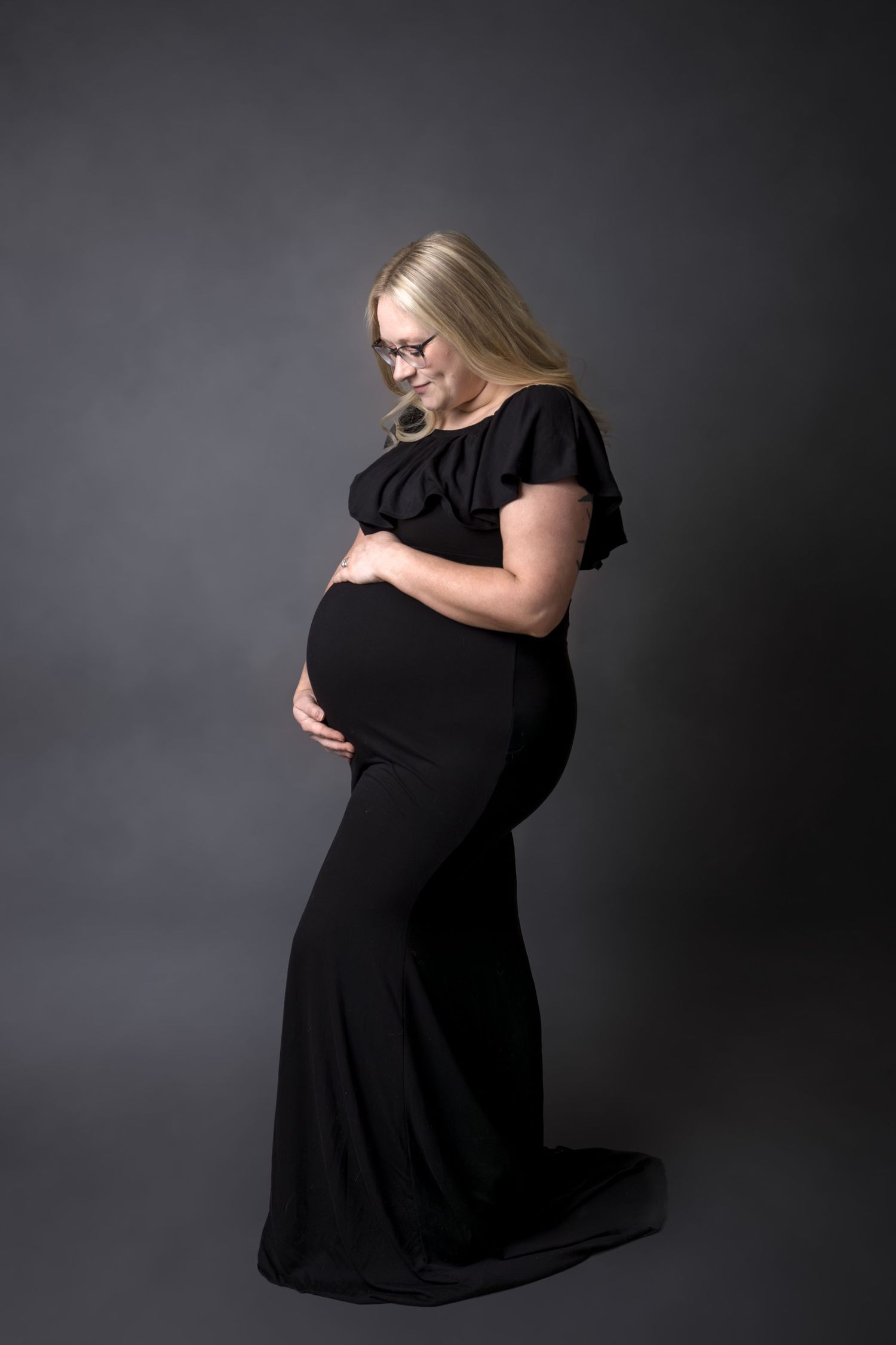 Pregnant woman in black maternity gown cradling baby bump, studio portrait with gray background.