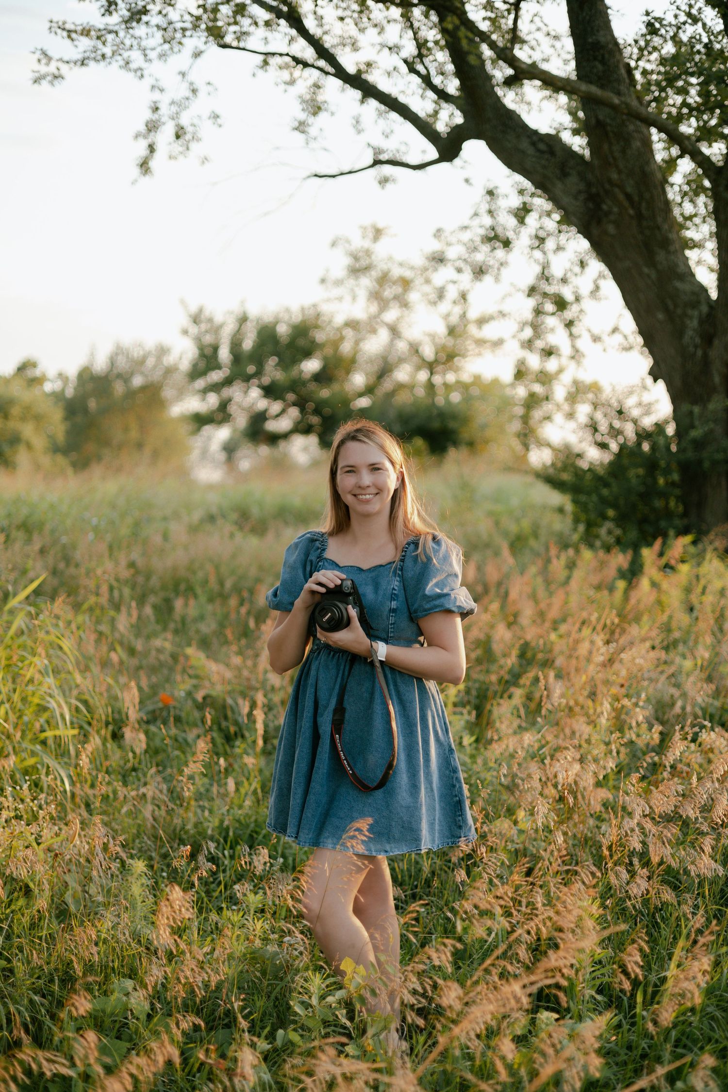 Young woman in blue dress holding camera standing in golden field near tree at sunset.