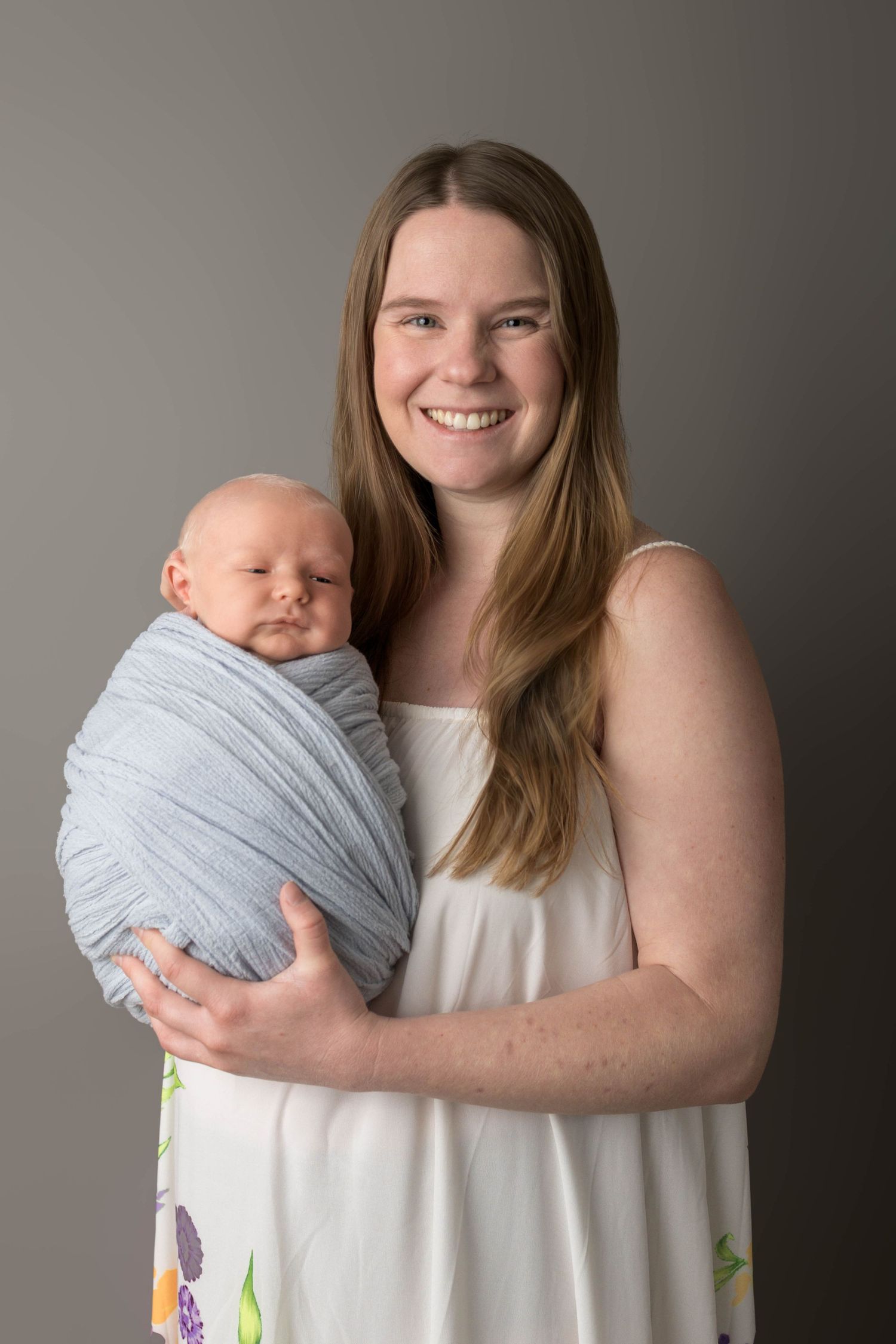 Smiling young mother in white dress holds sleeping newborn baby wrapped in blue swaddle against gray background.