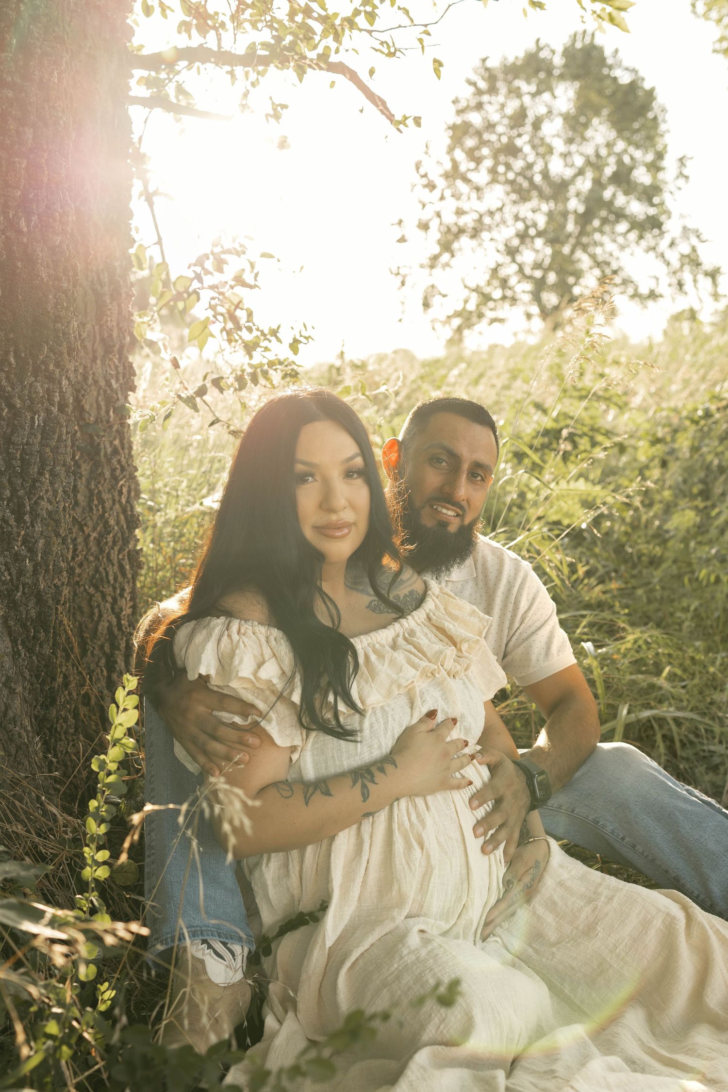 Couple sitting under a tree in golden sunlight, woman in white off-shoulder dress with tattoos, man in white shirt embracing her.
