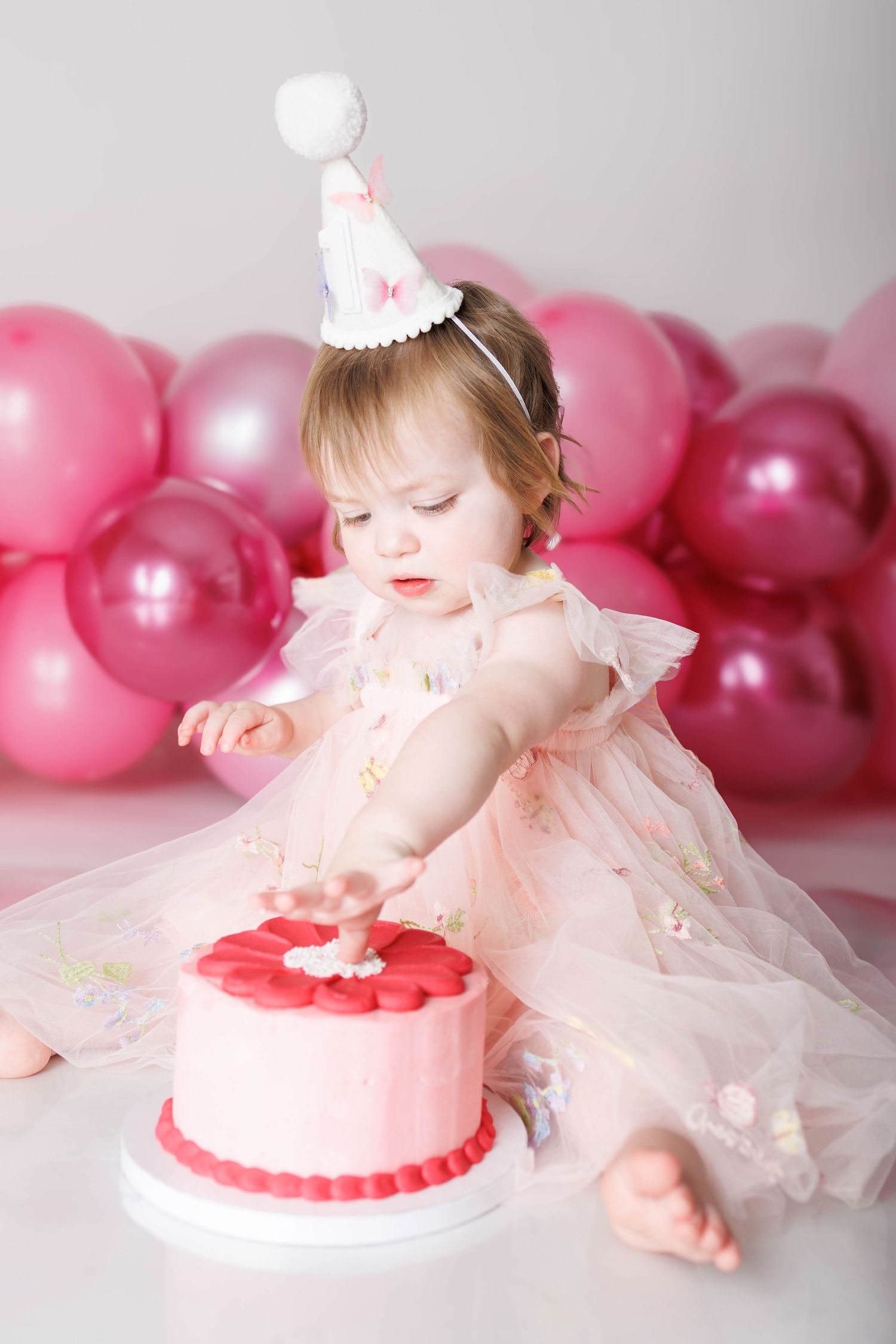 Baby girl in pink dress and party hat smashes her first birthday cake surrounded by pink balloons.