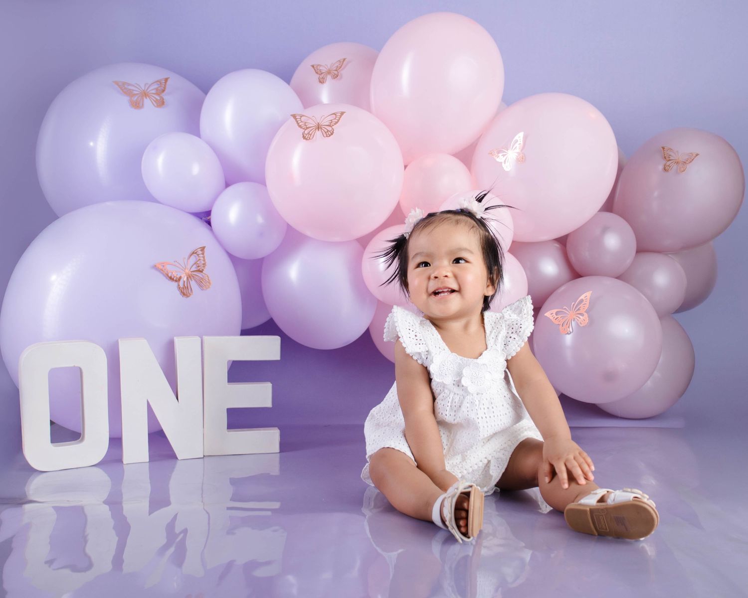 Baby wearing white dress sits among pink and purple balloons with butterfly decorations for first birthday celebration.