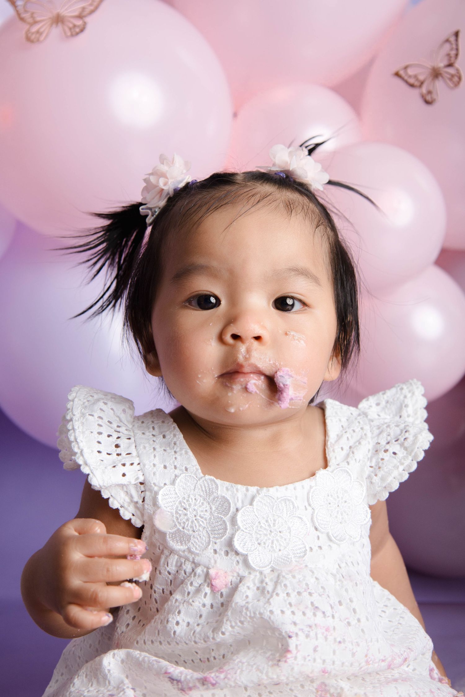 Sweet celebration scene with white eyelet dress against pink and purple balloon backdrop and butterfly decorations.