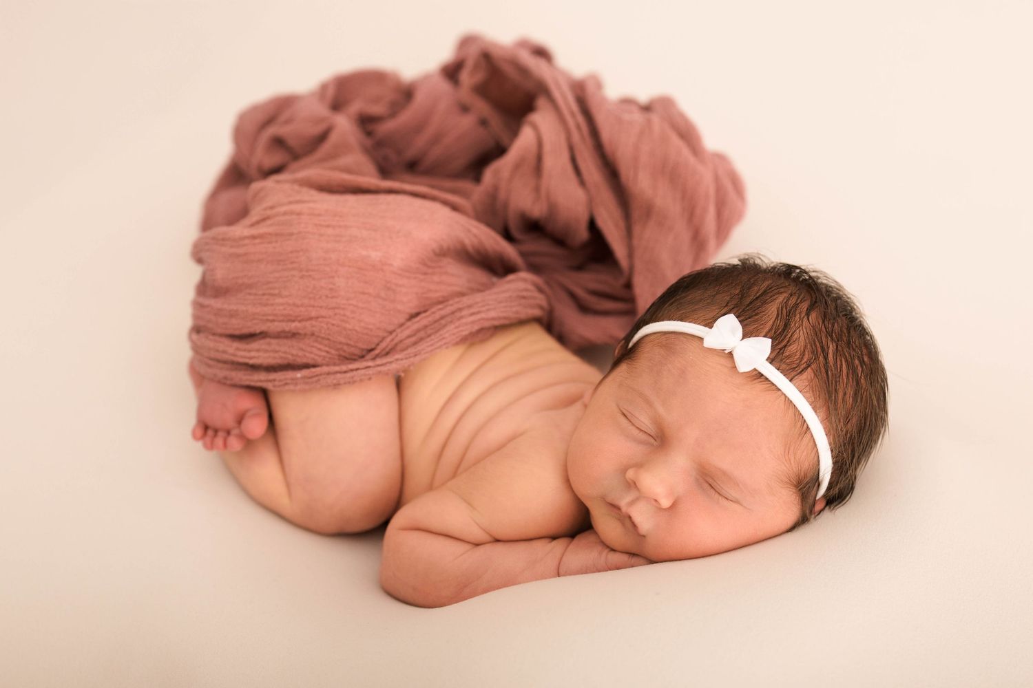 Peaceful newborn baby girl sleeping wrapped in a soft dusty rose pink blanket with a white bow headband.