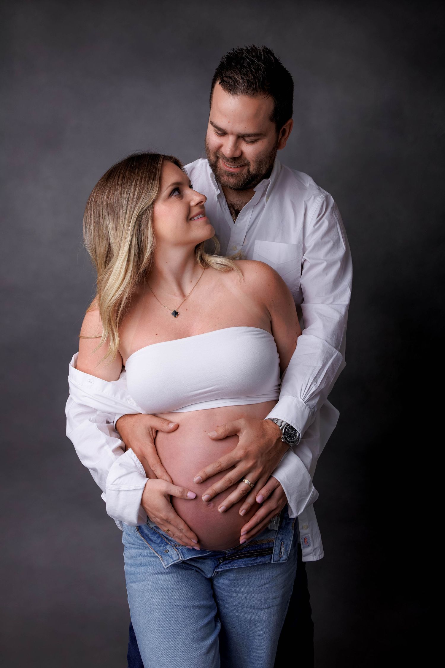 Couple embracing during maternity photoshoot, man holding pregnant woman's belly, wearing white tops and jeans on gray background.