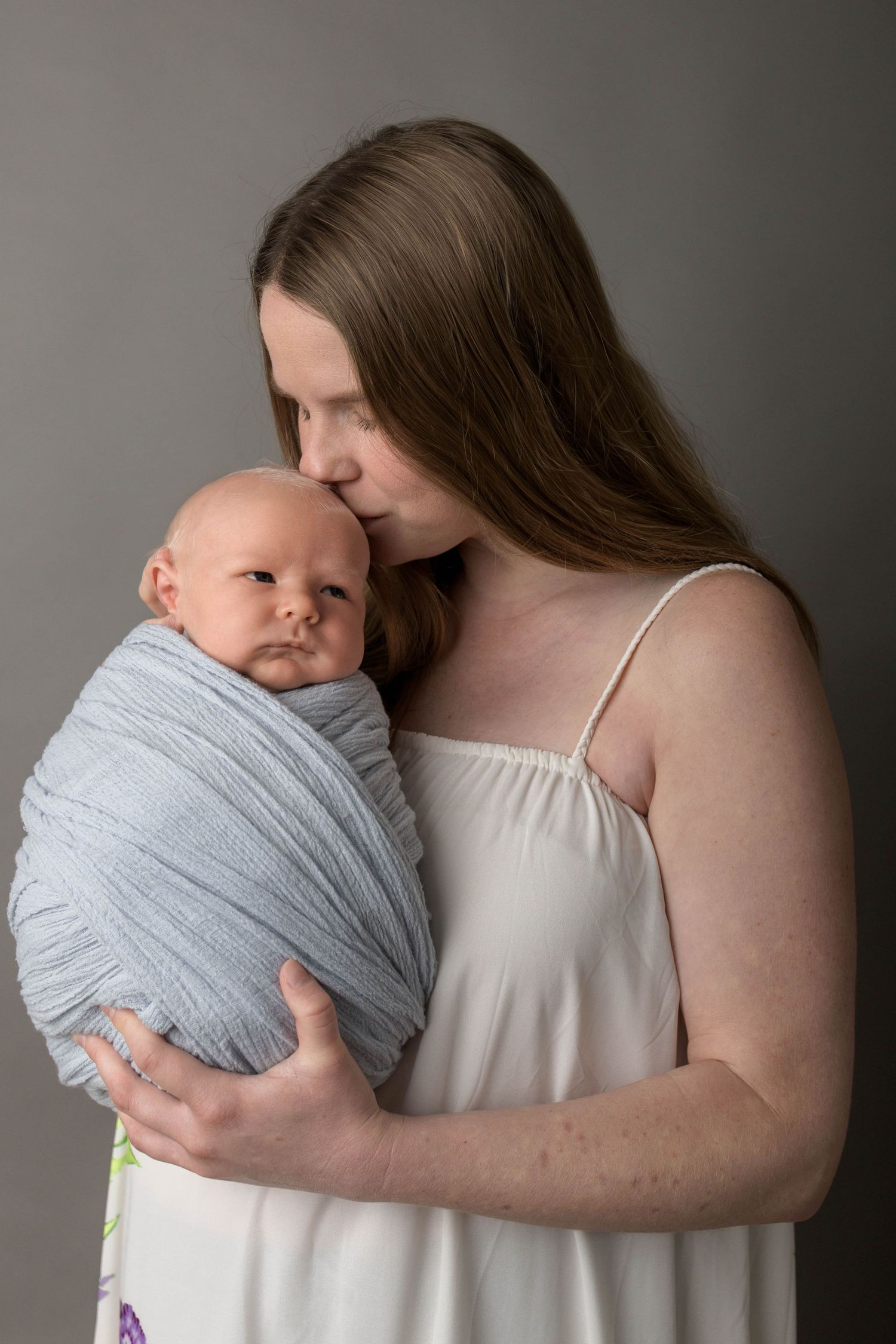 A mother in a white dress tenderly kisses her swaddled newborn baby wrapped in a blue cloth.