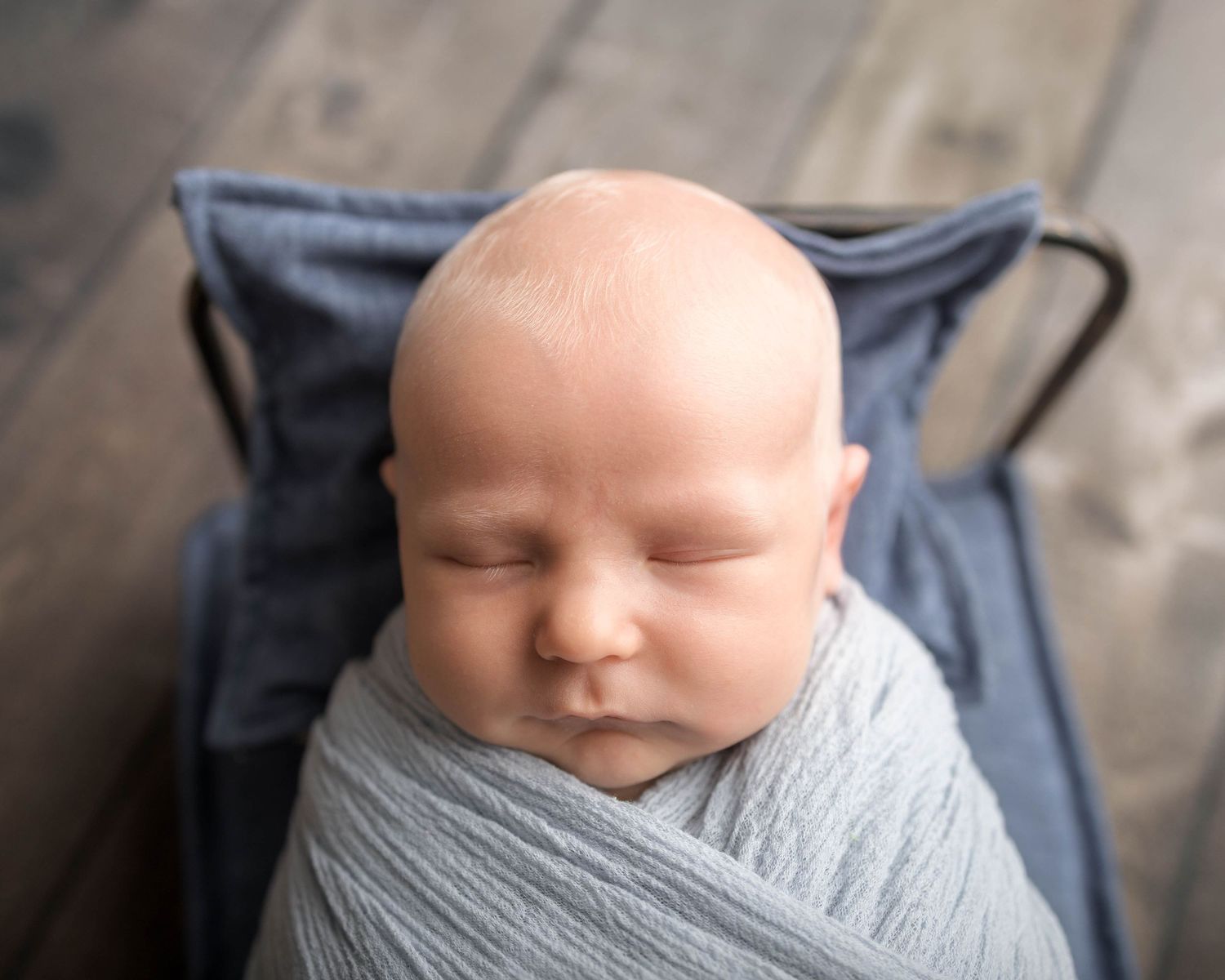Newborn baby swaddled in a soft gray wrap, sleeping peacefully in a small blue cot on a wooden floor.