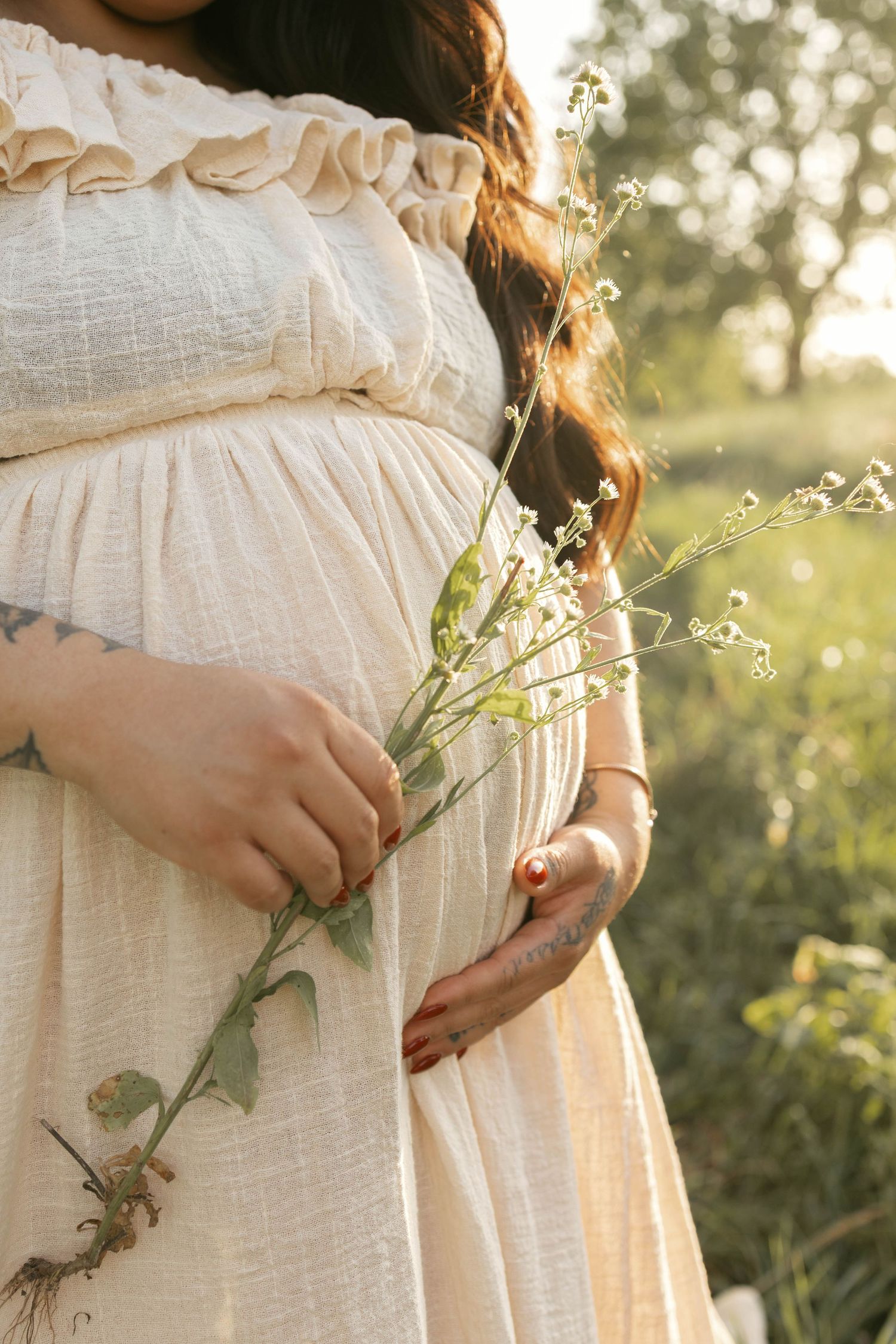 Close-up of pregnant woman in cream dress holding wildflowers in golden sunlight.