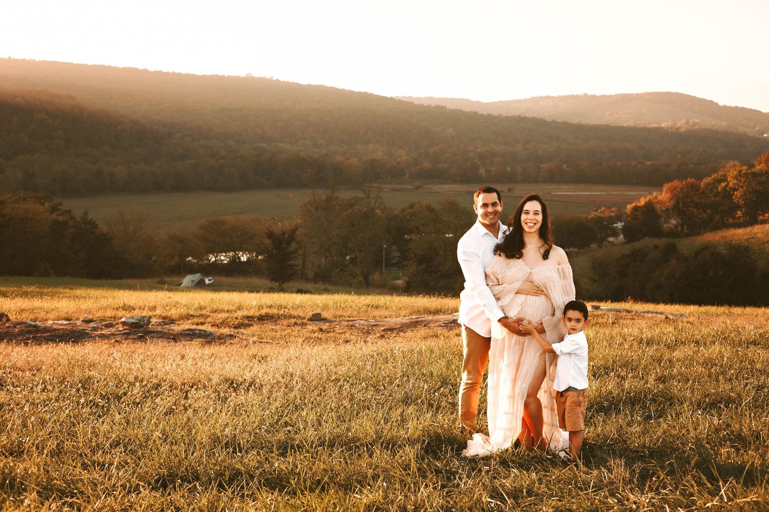 A family in white outfits sits together in a golden meadow at sunset with rolling hills in the background.