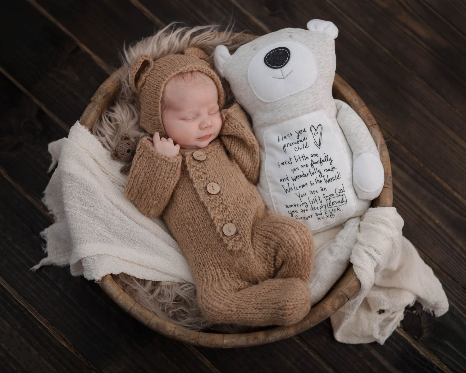 Newborn photography set with brown knit sweater, soft white blankets, and plush teddy bear on rustic wooden floor.