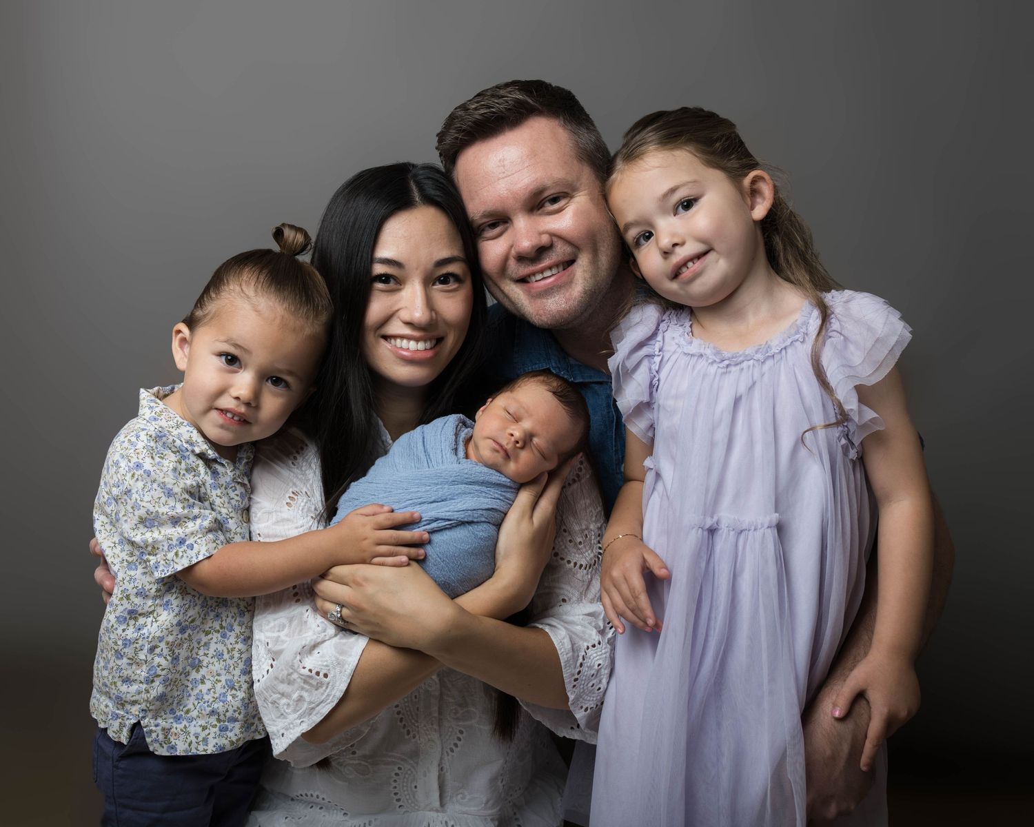 Happy family of five posing for a newborn portrait, parents with two toddlers and a sleeping baby wrapped in blue.