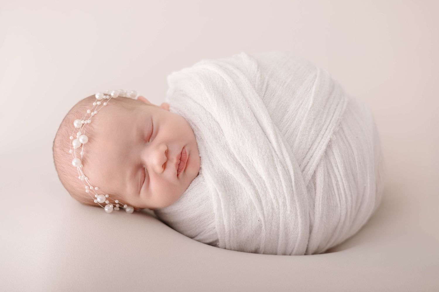 Sleeping newborn baby swaddled in white wrap with floral headband on neutral background.