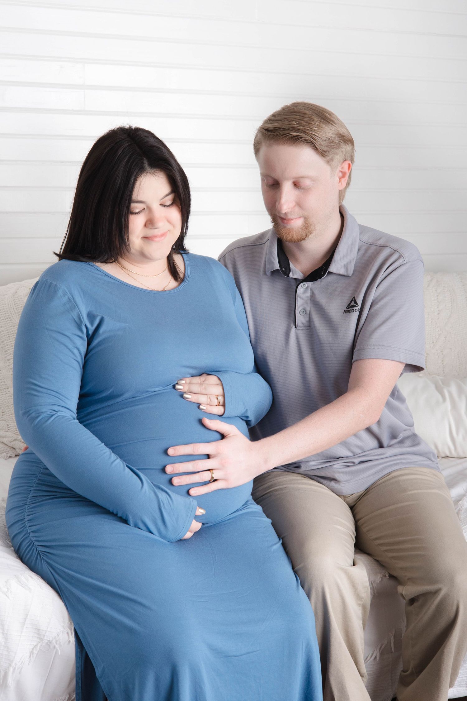 Expecting parents share a tender moment on a white couch as they lovingly touch the pregnant belly.