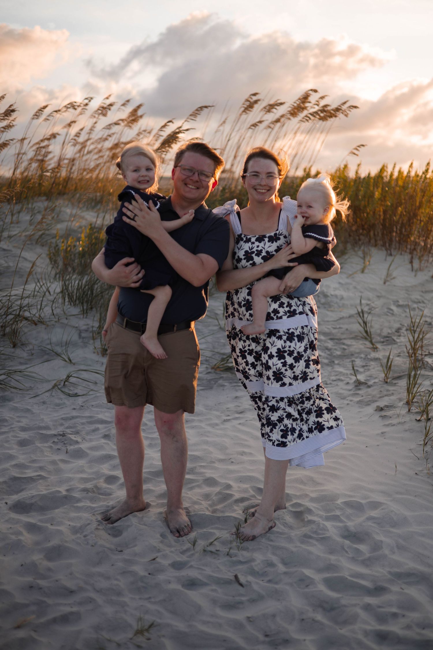 Group poses casually on beach sand dunes with golden sunset lighting and sea grass.