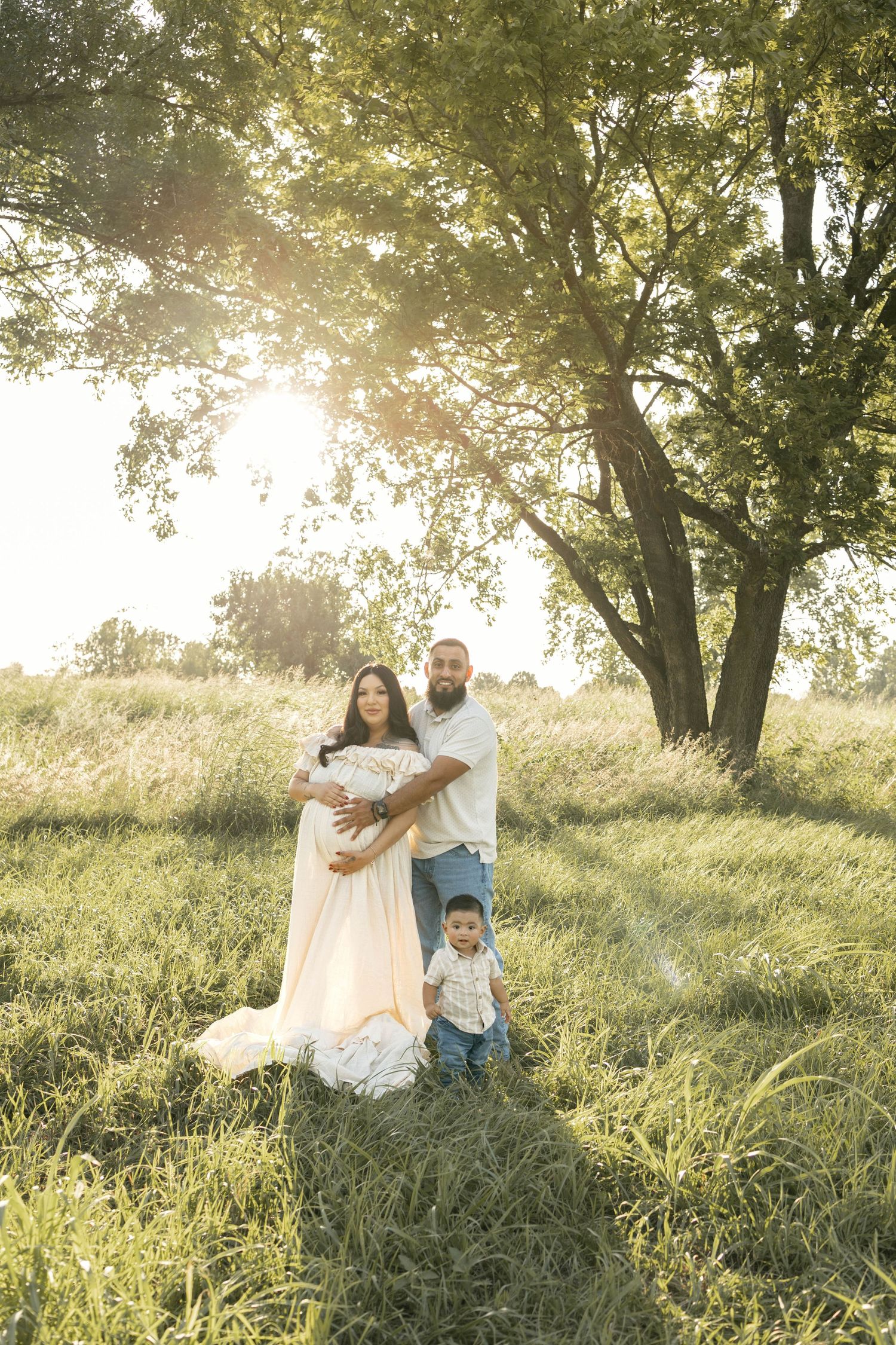 Pregnant woman in peach gown with husband and toddler son posing in sunlit green field near large tree.