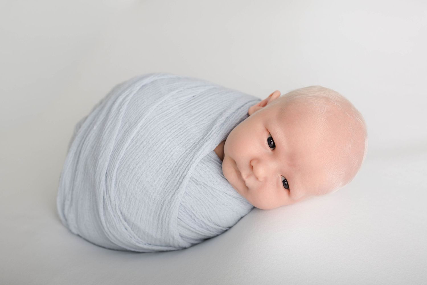 Newborn baby swaddled in a soft blue muslin wrap, lying peacefully on a white background.