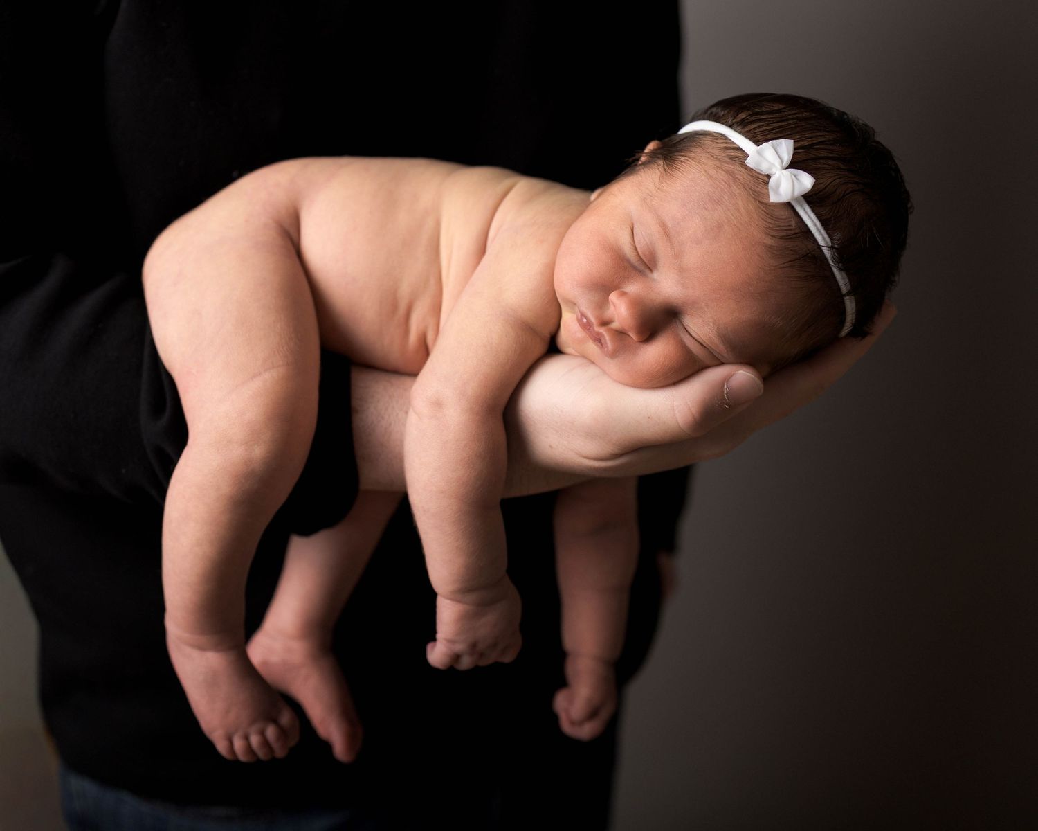Newborn baby girl sleeping peacefully on parent's hand, wearing white bow headband, in professional studio portrait.