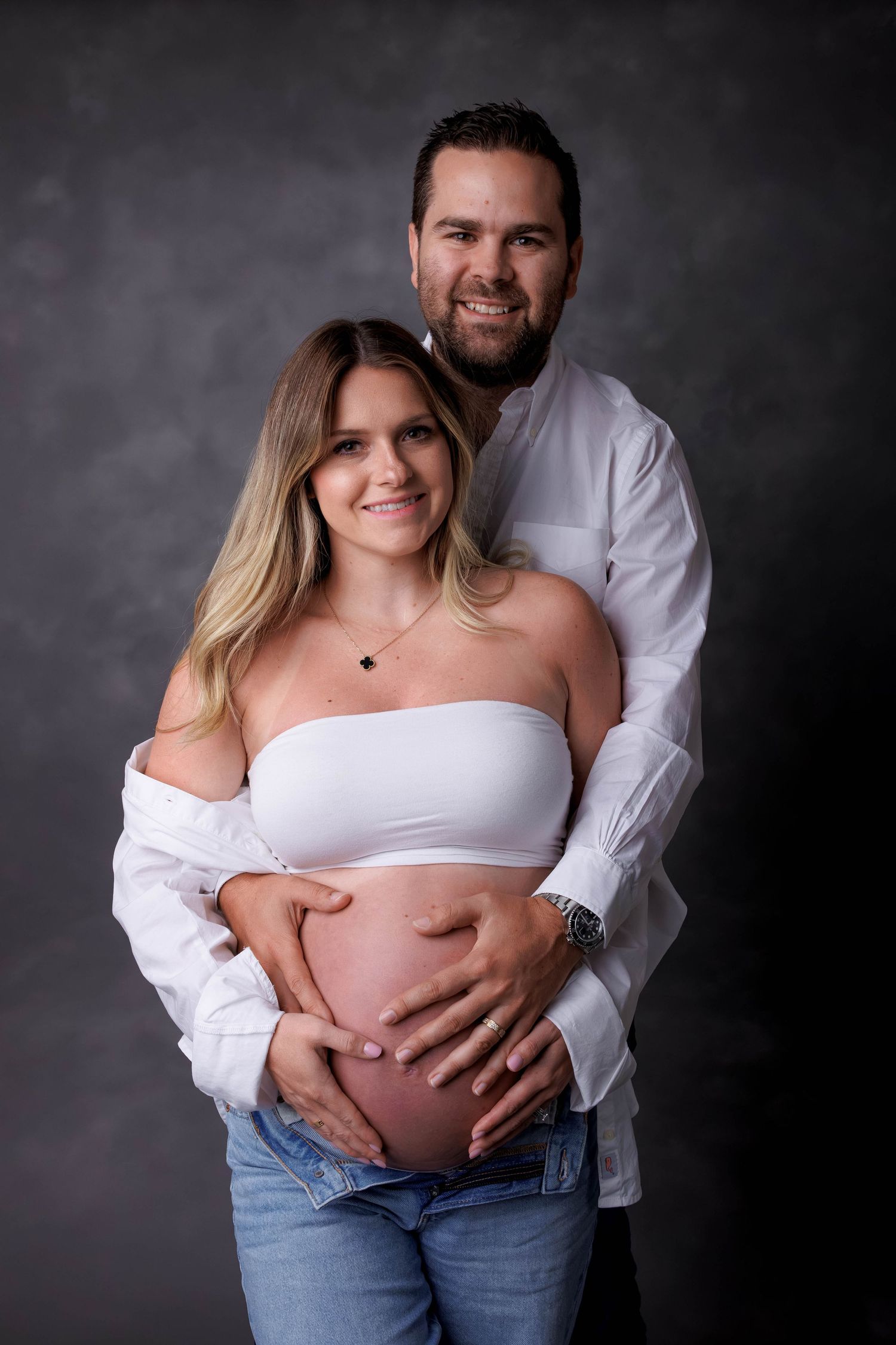Couple posing for maternity photo shoot, woman in white tube top showing baby bump, man embracing her from behind in studio.