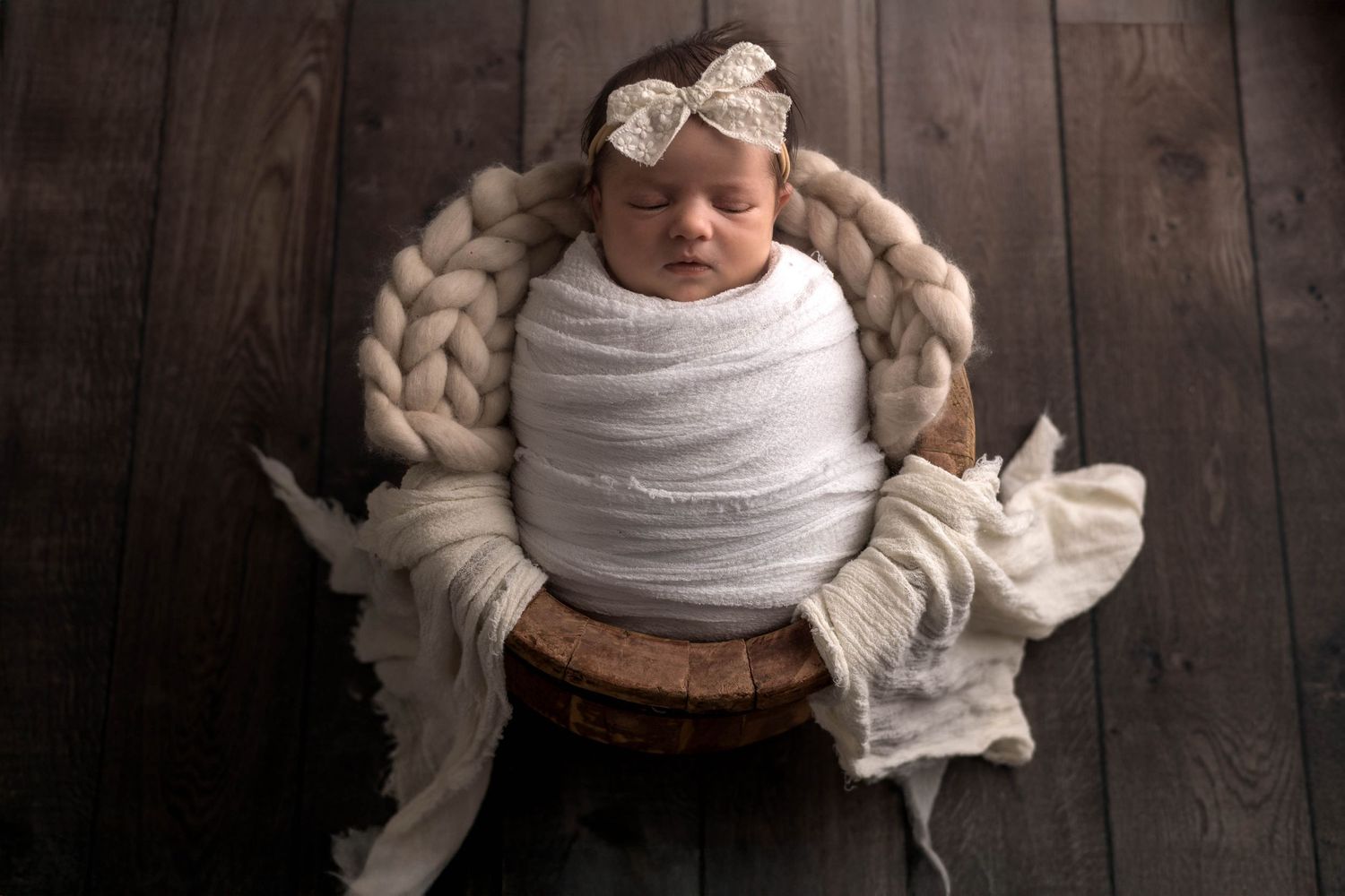A newborn baby wrapped in white fabric lies on a dark wooden floor during a professional photo session.