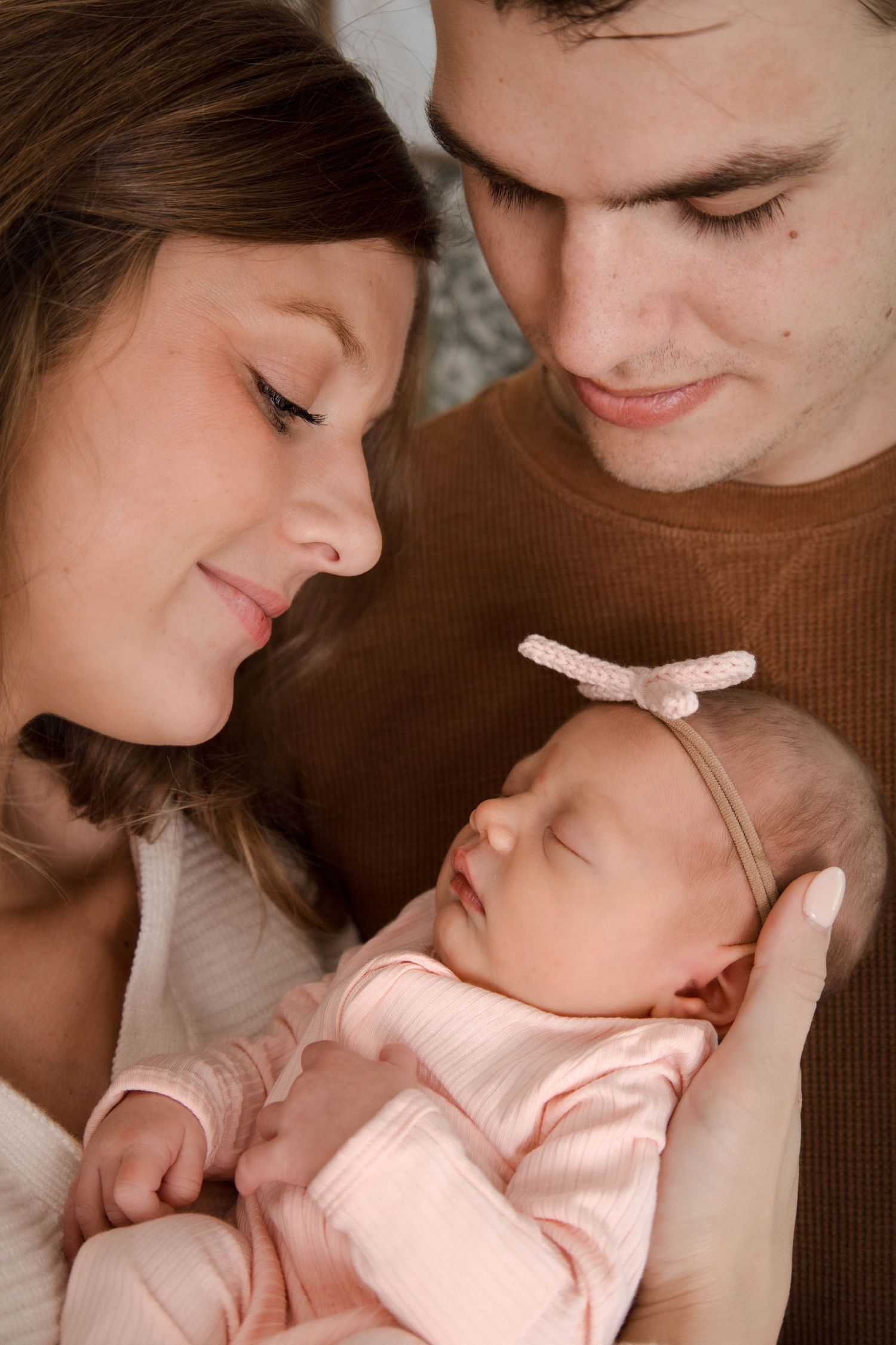 Tender family moment as parents share intimate bonding time with sleeping newborn in soft pink outfit.